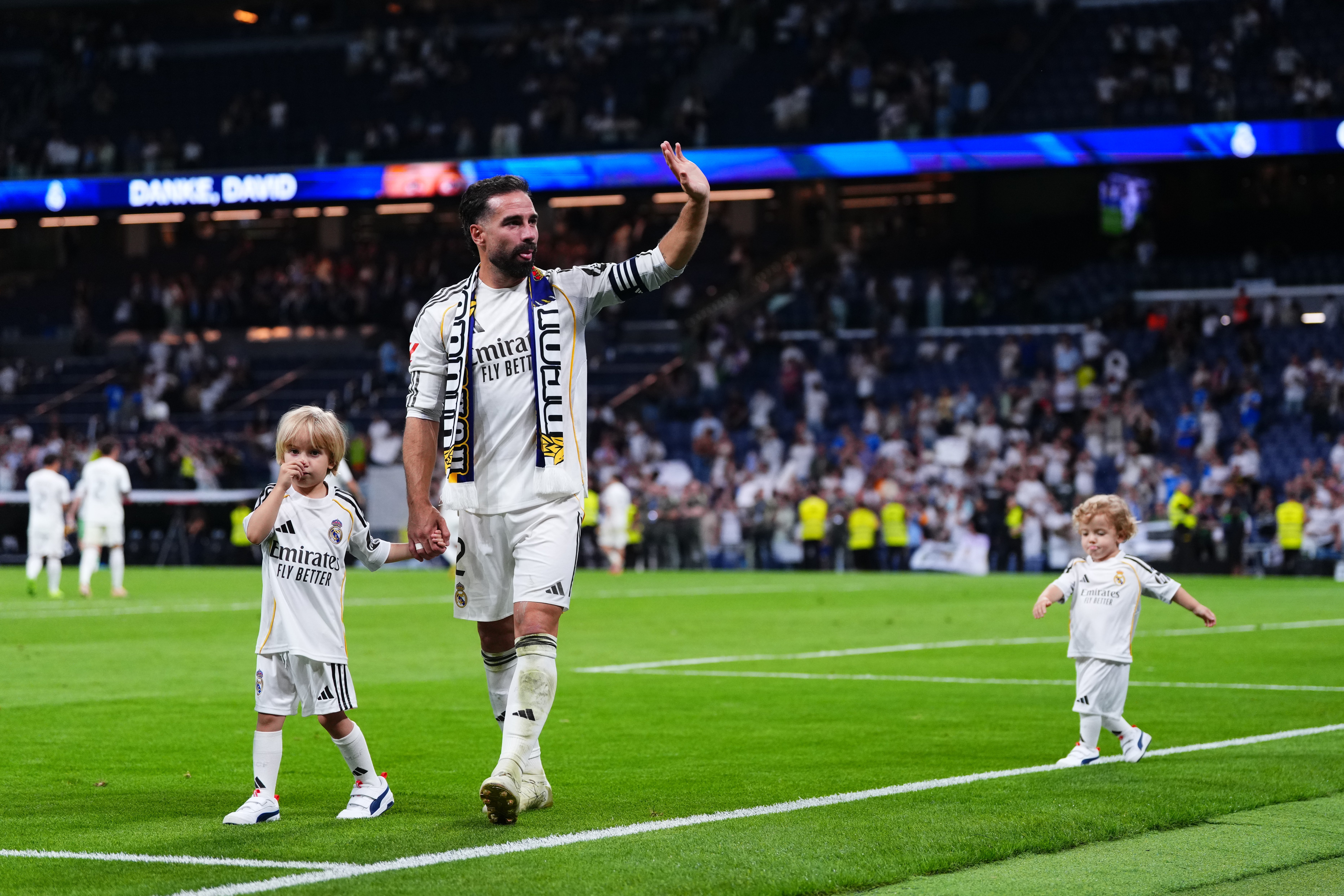 En fotbollsspelare i vit Real Madrid-tröja går på fotbollsplanen tillsammans med två små barn, också i vita tröjor. Spelaren viftar till publiken under en match.