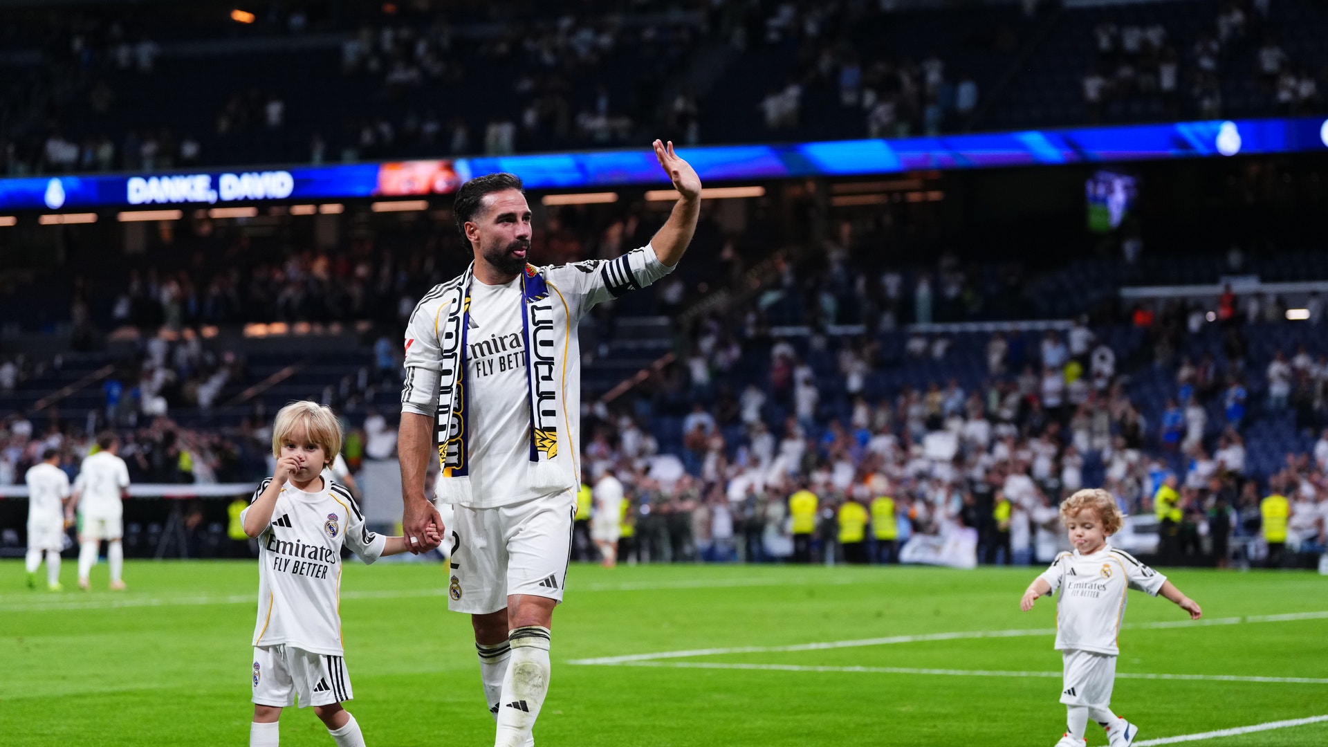 En fotbollsspelare i vit Real Madrid-tröja går på fotbollsplanen tillsammans med två små barn, också i vita tröjor. Spelaren viftar till publiken under en match.
