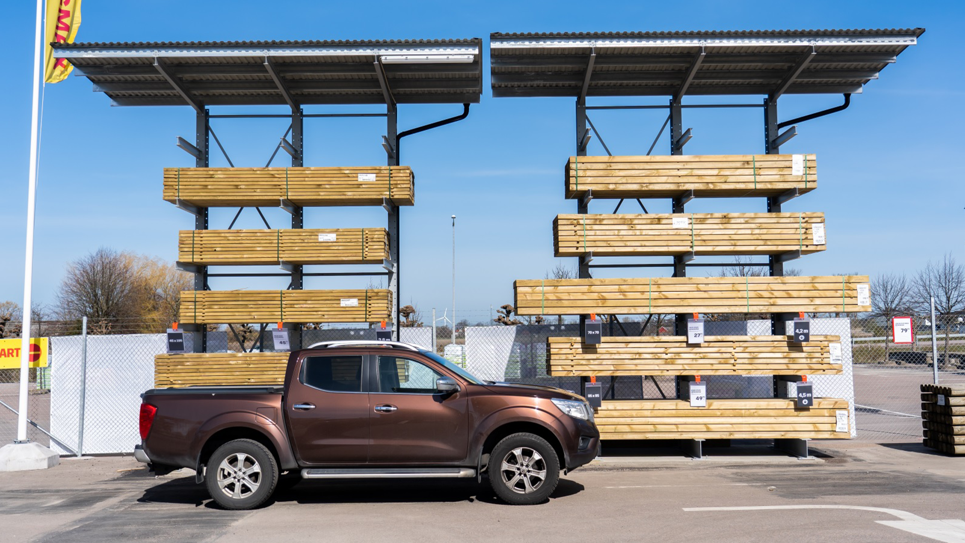 En brun pickup-truck parkerad framför en Byggmax-butik med en stor utomhusutställning av trälämningar och byggmaterial på höga ställningar.