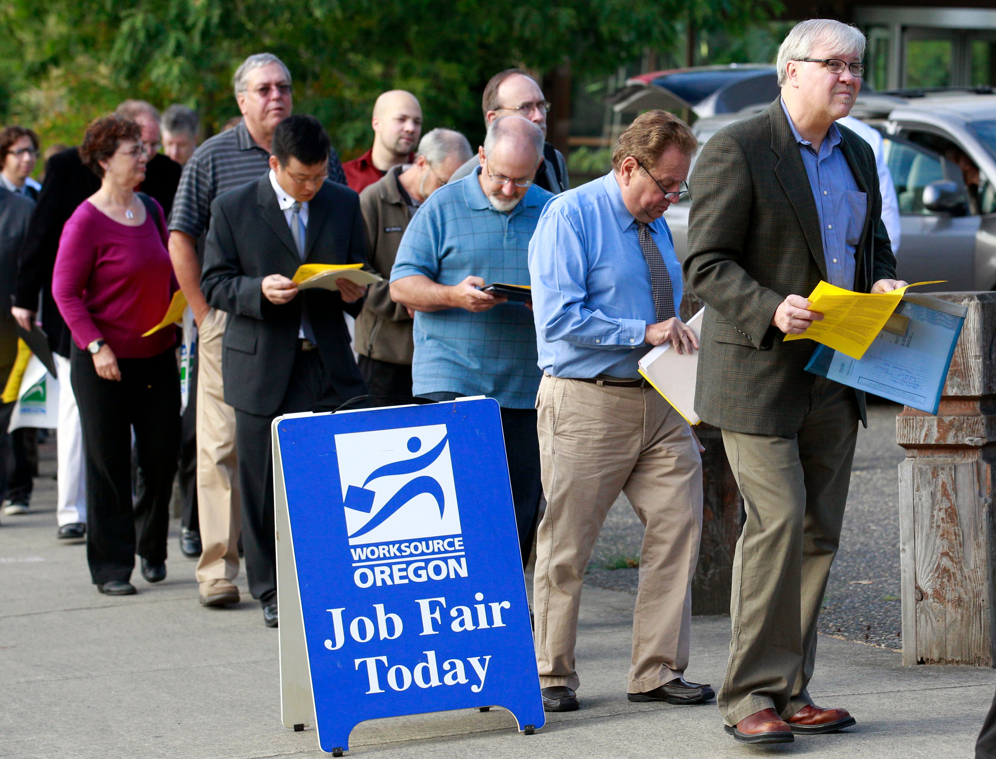 En grupp människor står i kö vid en jobbmässa arrangerad av WorkSource Oregon. Många håller gula papper i händerna. En blå skylt framför säger 'Job Fair Today'.