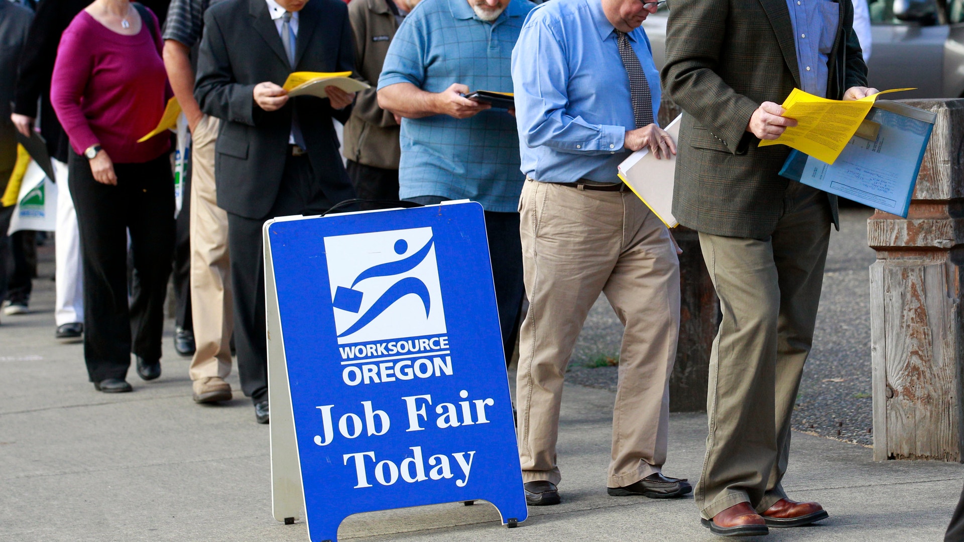En grupp människor står i kö vid en jobbmässa arrangerad av WorkSource Oregon. Många håller gula papper i händerna. En blå skylt framför säger 'Job Fair Today'.