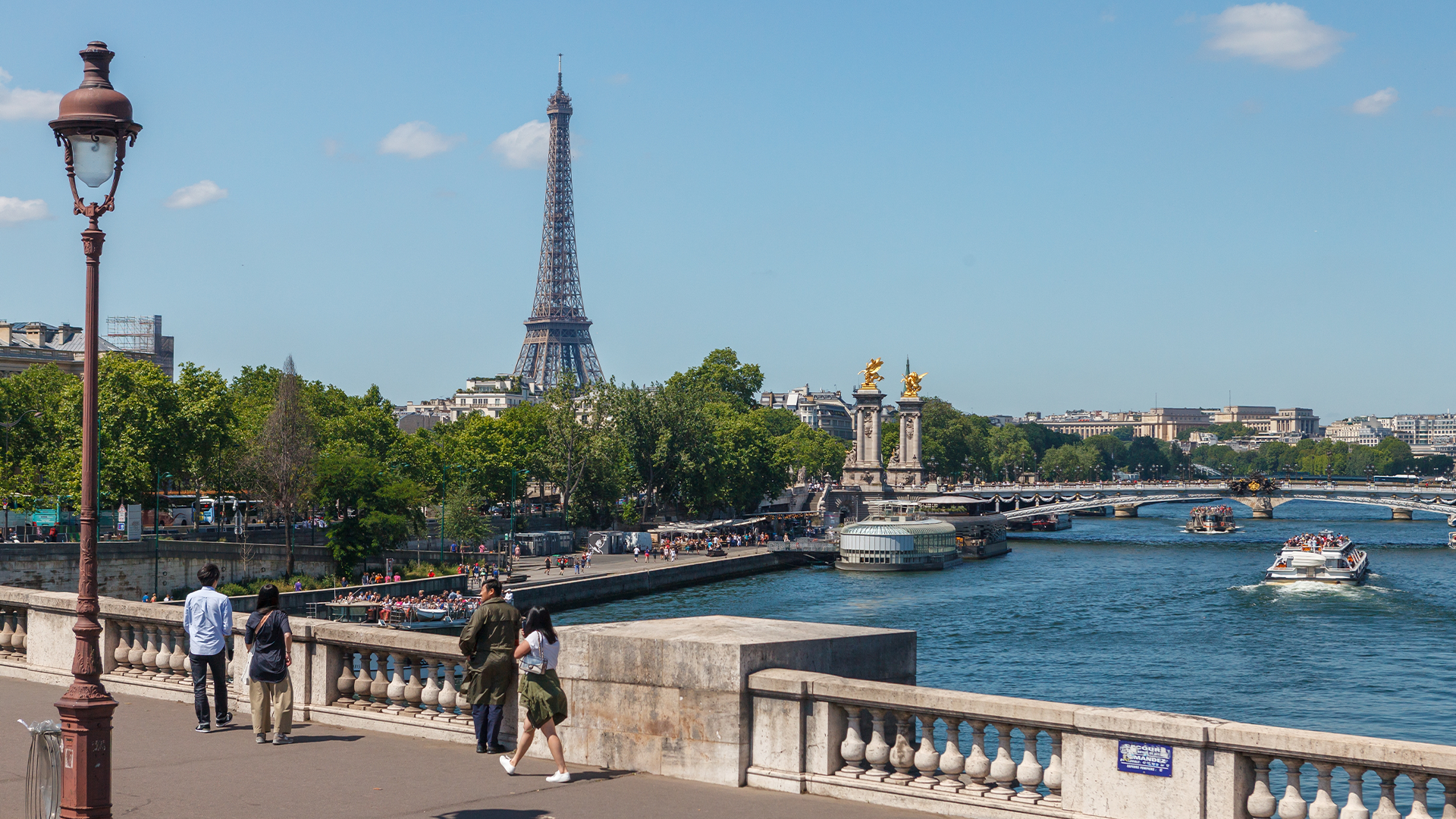 Utsikt över Seinen i Paris med Eiffeltornet i bakgrunden, båtar på vattnet och människor som promenerar på en bro.