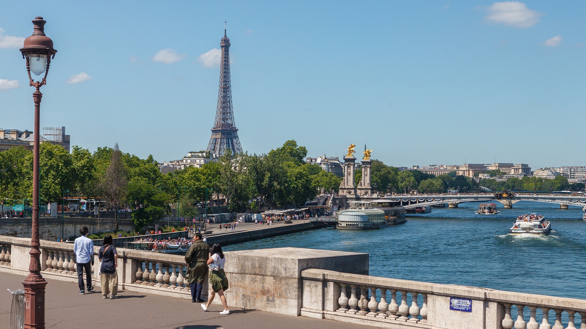 Utsikt över Seinen i Paris med Eiffeltornet i bakgrunden, båtar på vattnet och människor som promenerar på en bro.