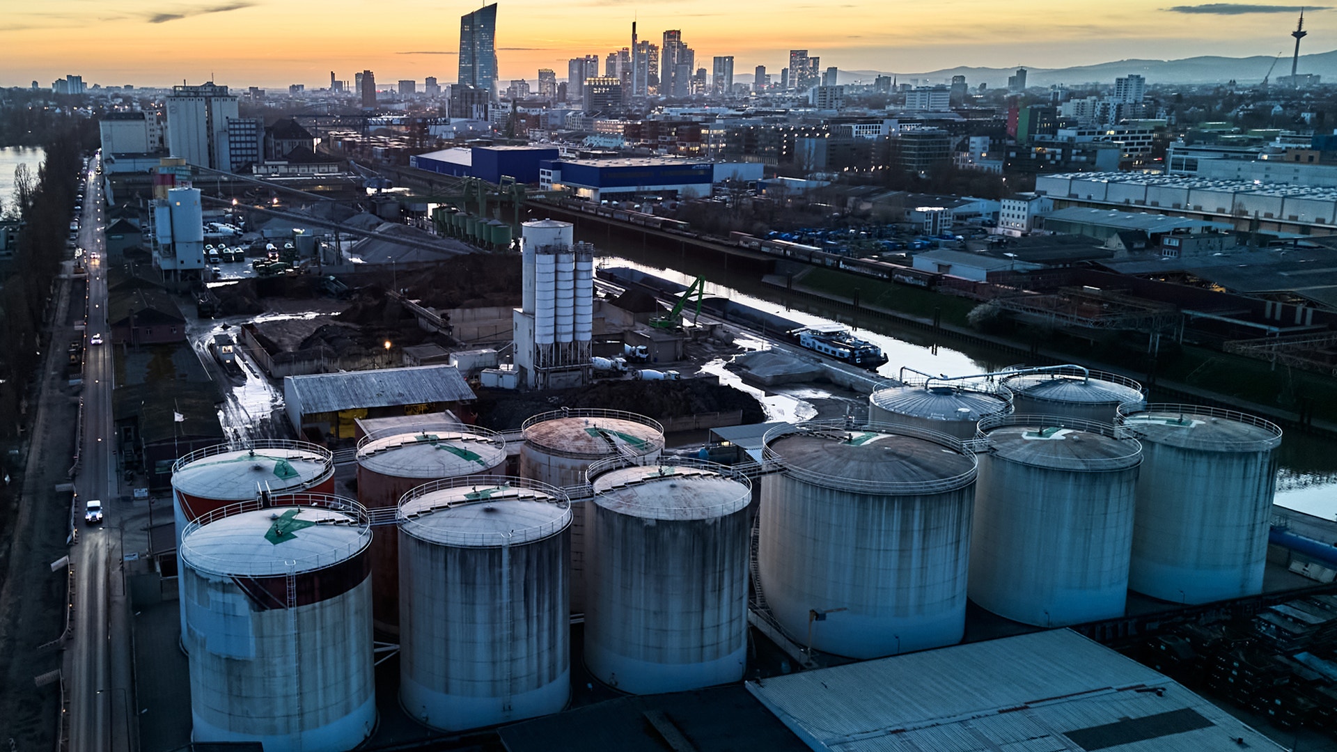 Industriell anläggning med stora lagertankar i förgrunden och stadshorisont i bakgrunden vid solnedgång.