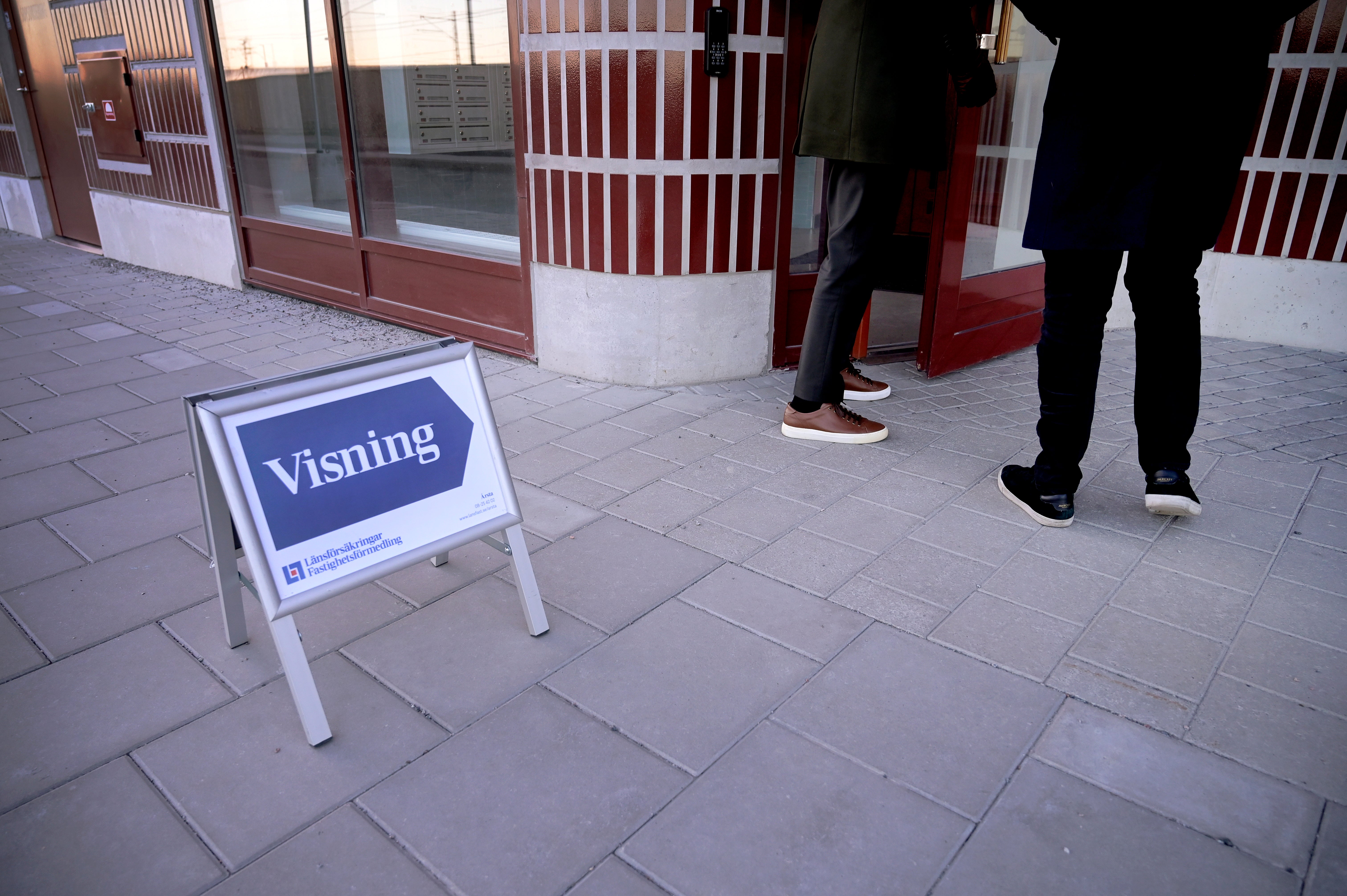 Ett vitt ställ med blått skylt märkt 'visning' står på stenlagd gata framför en butiksentré med röda och vita dörrar. Två personer står vid ingången.
