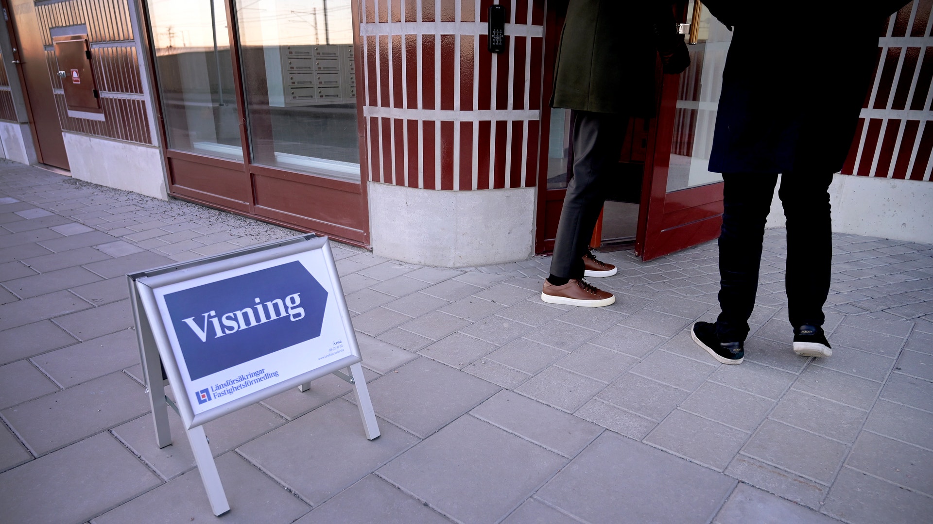 Ett vitt ställ med blått skylt märkt 'visning' står på stenlagd gata framför en butiksentré med röda och vita dörrar. Två personer står vid ingången.