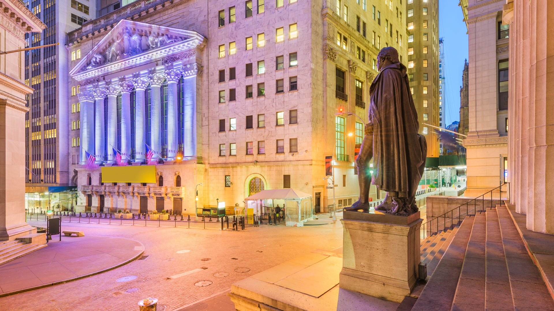 Nattlig vy av New York Stock Exchange-byggnaden med klassisk arkitektur, omgiven av höga byggnader och en staty i förgrunden.