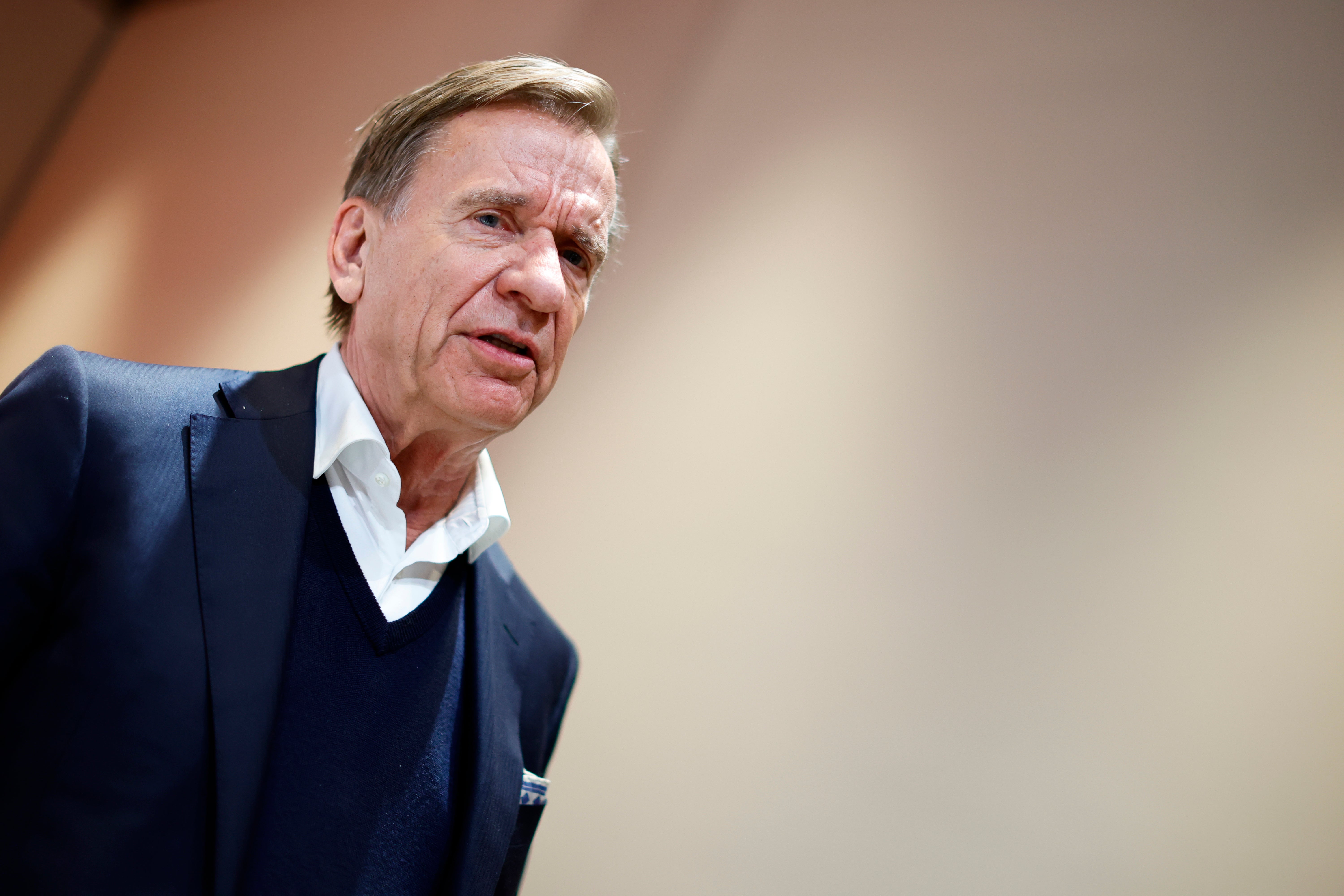Porträtt av en man i mörkblå tröja och vit skjorta mot neutral bakgrund.