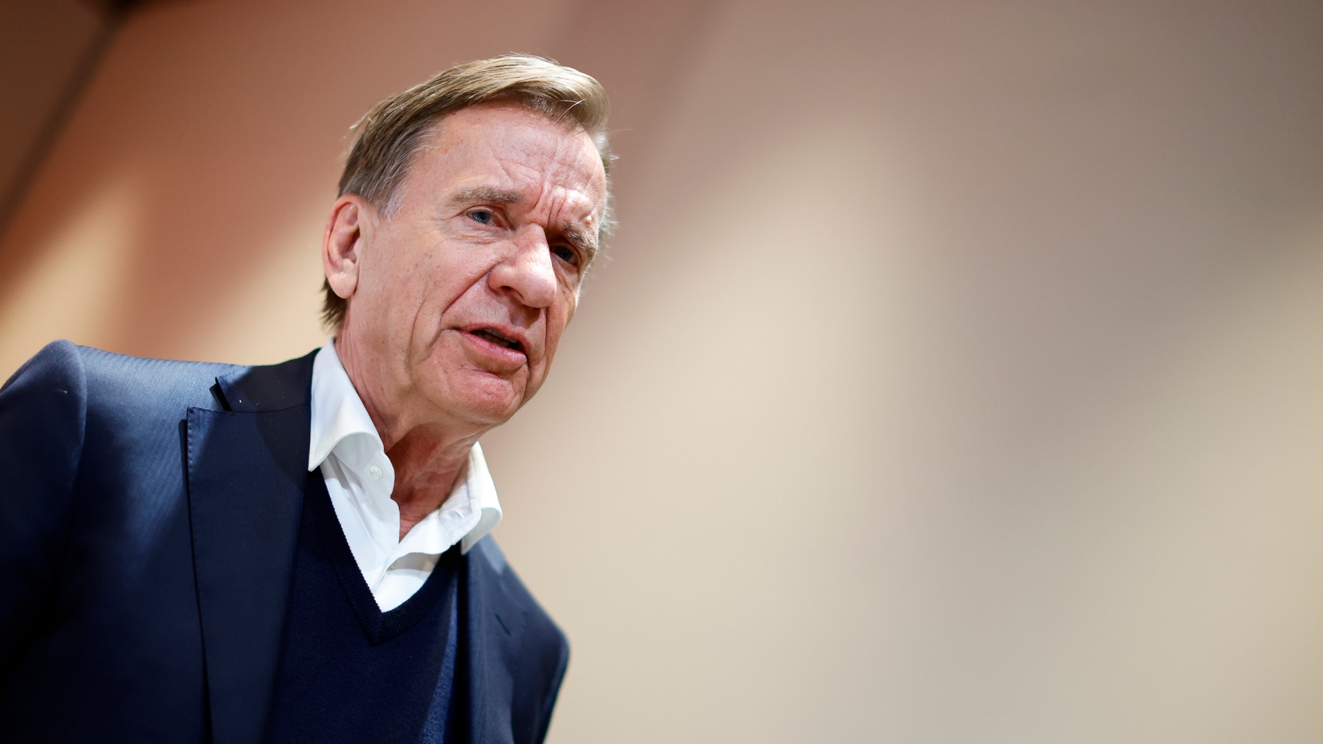 Porträtt av en man i mörkblå tröja och vit skjorta mot neutral bakgrund.