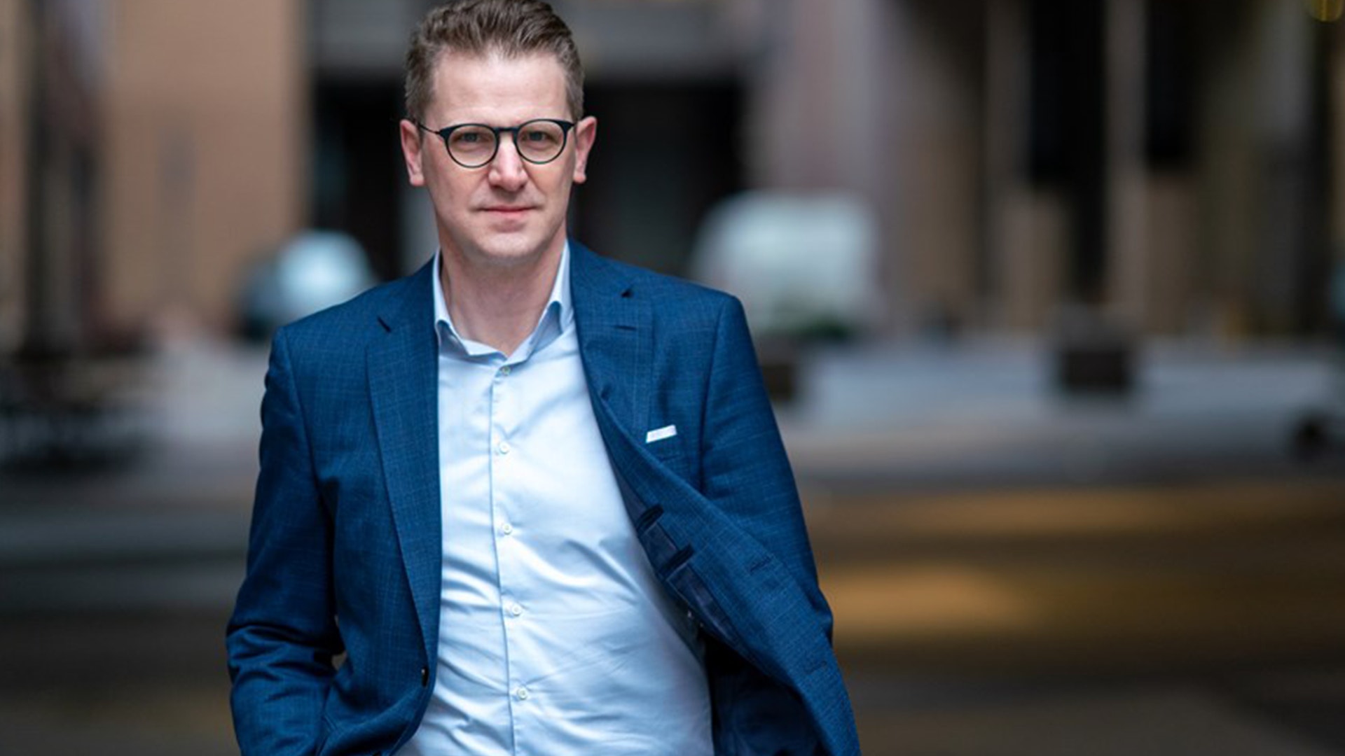 Porträtt av en man i blå kavaj och ljus skjorta som står framför en suddig stadsmiljö.