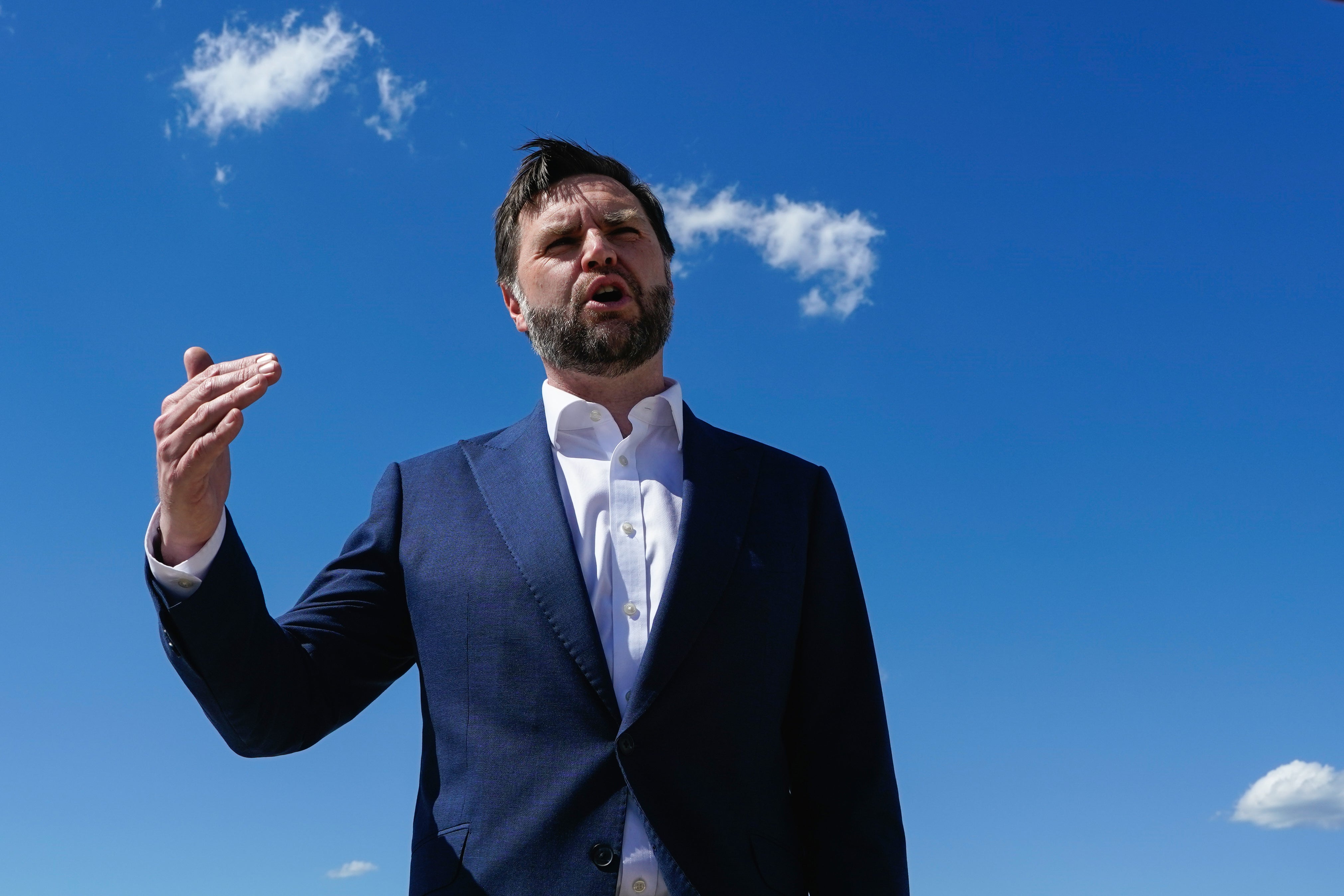 Man i mörkblå kostym och vit skjorta som gestikulerar mot en klar blå himmel med vita moln.