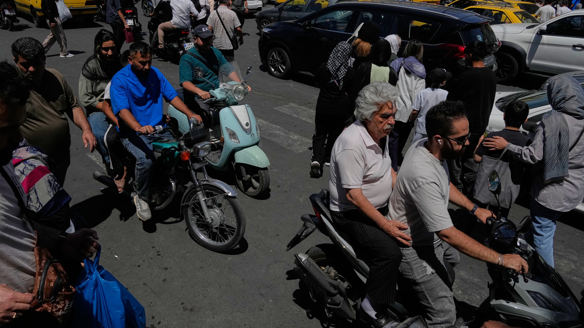 En gatuscen med flera personer och motorcyklar parkerade på en väg. Folk står och samlas runt fordon, några bär blå kläder. Det är dagtid och solen skiner på asfalten.