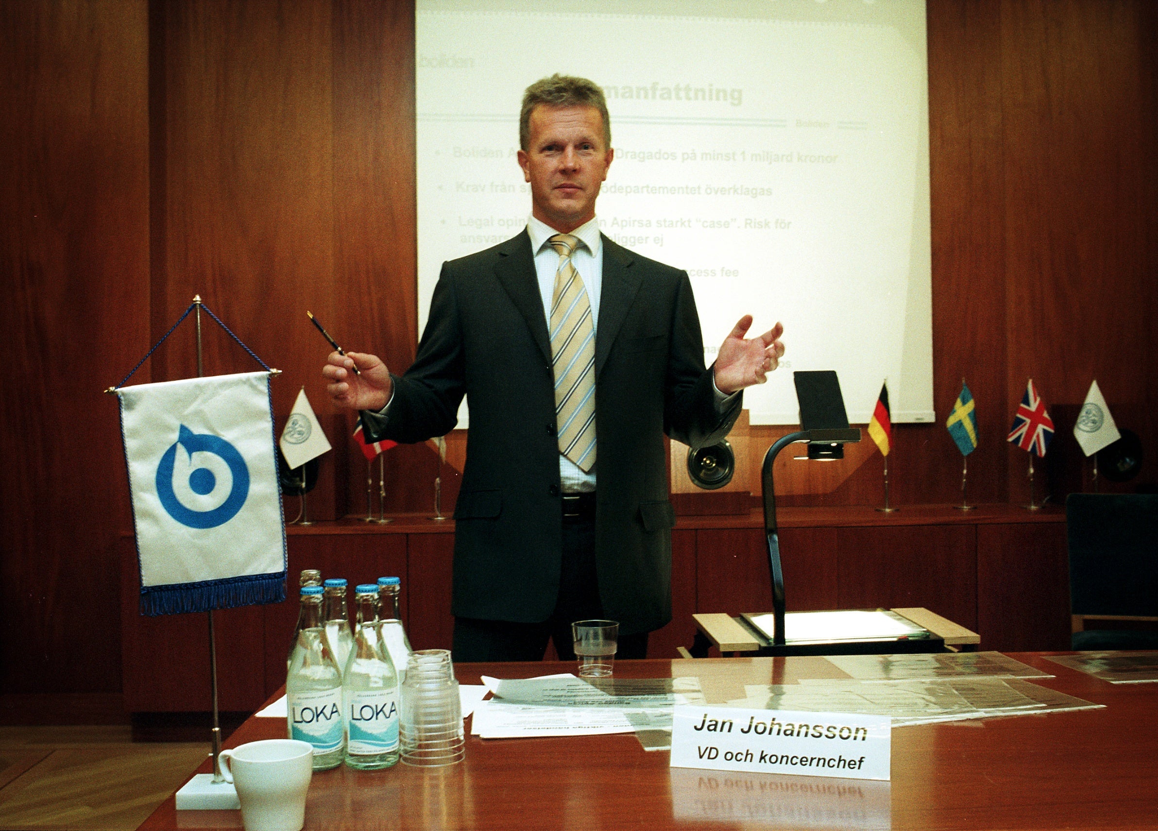 Man i mörk kostym och slips presenterar vid ett podium under en konferens. Han gestikulerar medan han talar. På bordet framför honom ligger flaggor, vattenflaskor och en namnbricka.