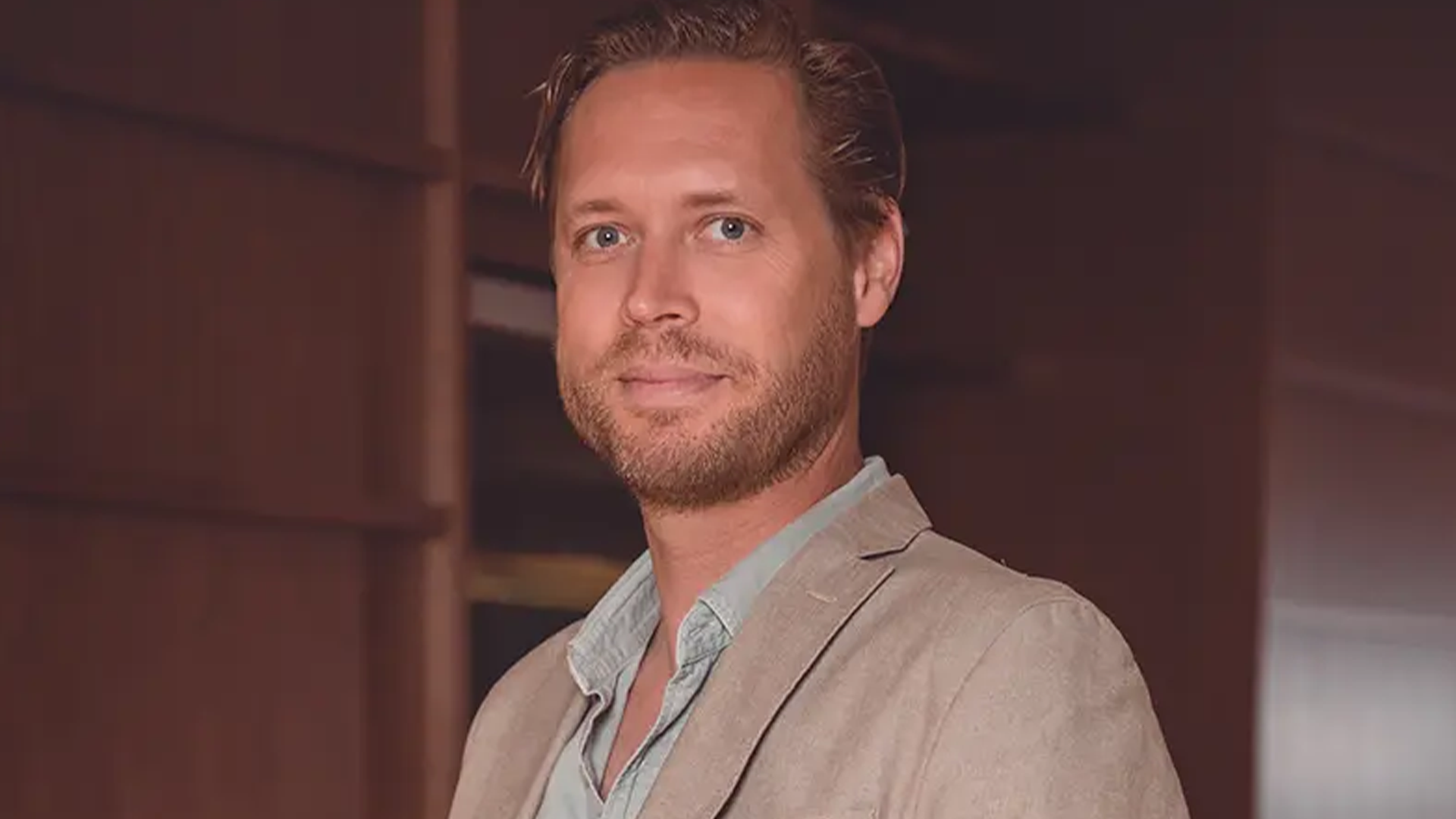 Oscar Stibeck, Fastator, en leende man med kort skägg i beige kavaj och ljusblå skjorta.