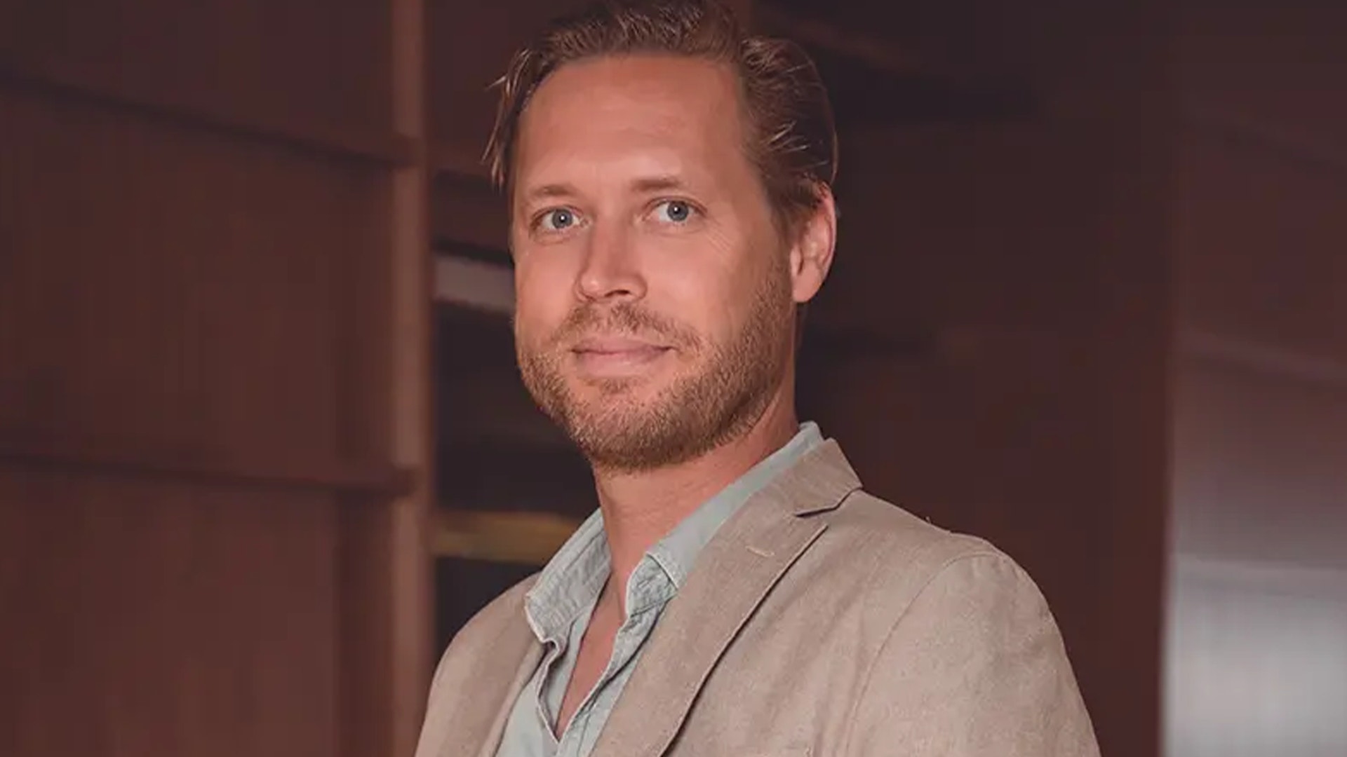 Oscar Stibeck, Fastator, en leende man med kort skägg i beige kavaj och ljusblå skjorta.