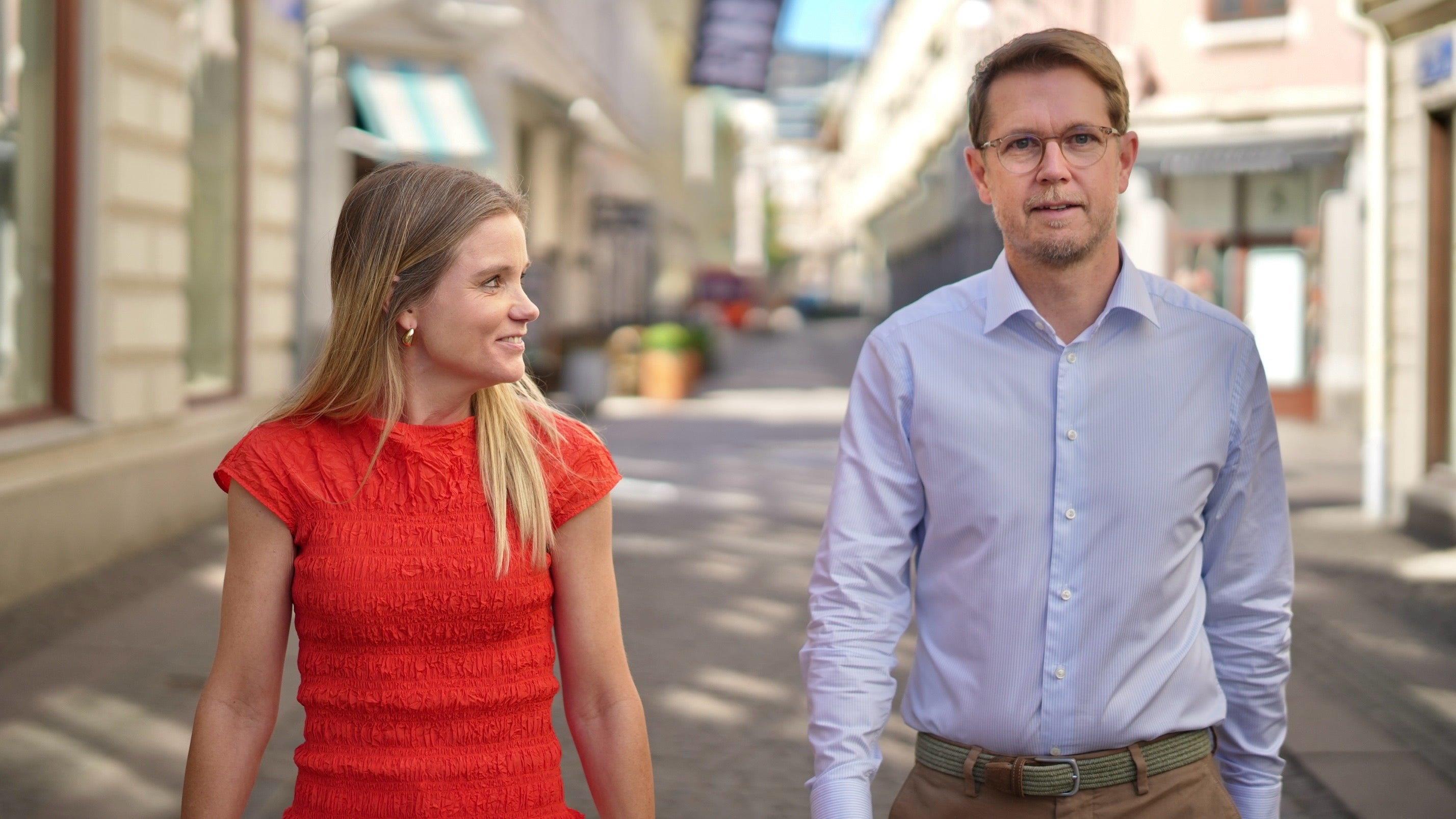 En kvinna i röd klänning och en man i ljusblå skjorta går tillsammans på en solig stadsgata.
