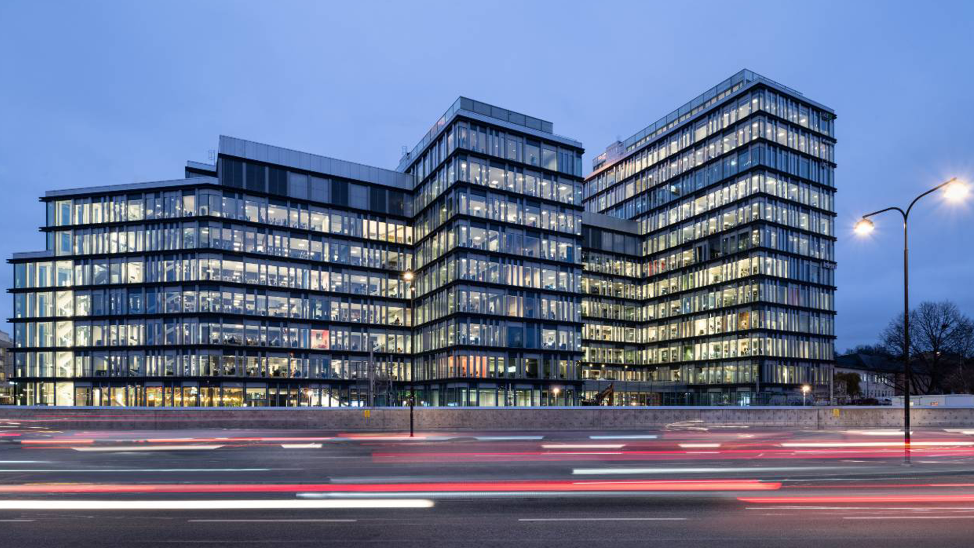 Modern kontorsbyggnad fotograferad på kvällen med upplysta fönster. Flera sammankopplade byggnadsdelar i glas och stål. Trafikljus från fordon syns framför byggnaden.