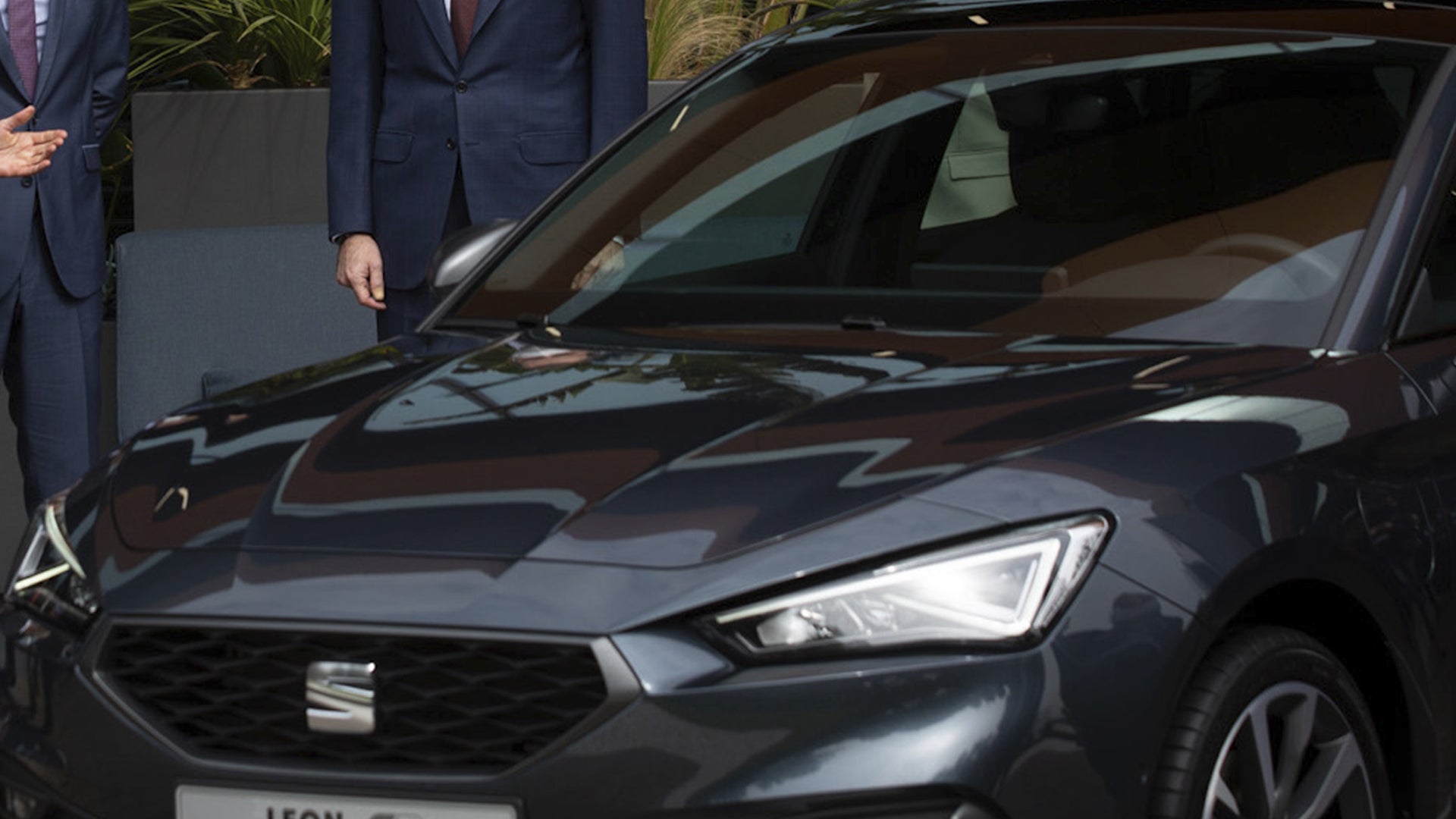 En mörk Seat Leon fotograferad snett framifrån, med två kostymklädda män stående bakom bilen vid en presentation.