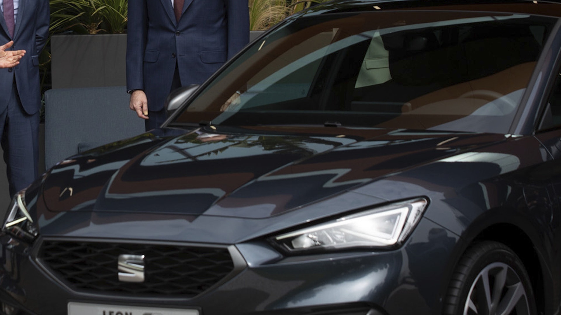 En mörk Seat Leon fotograferad snett framifrån, med två kostymklädda män stående bakom bilen vid en presentation.