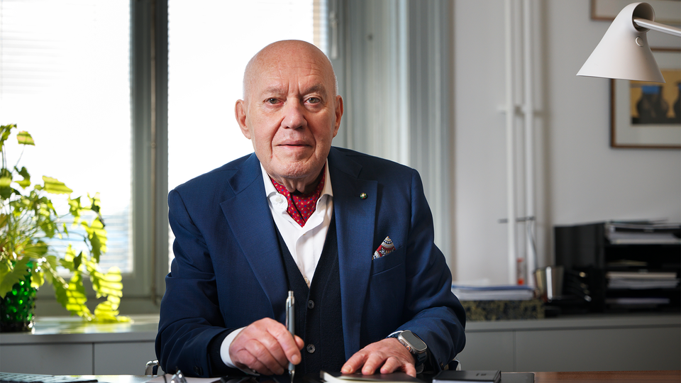 Porträtt av en äldre man i blå kostym och röd fluga sittande vid ett skrivbord på ett kontor.