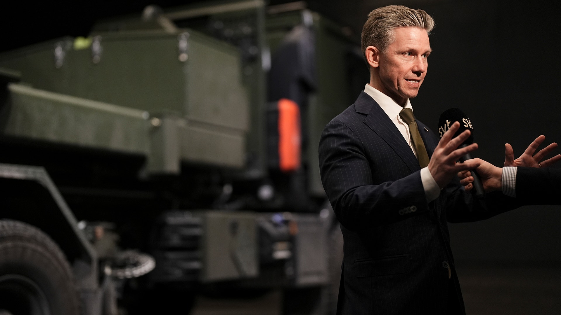 Pål Jonsson, Sveriges försvarsminister, håller ett tal och gestikulerar medan han talar. Han är klädd i en mörkblå kostym och ljus skjorta. I bakgrunden syns militär utrustning inklusive ett militärt fordon.