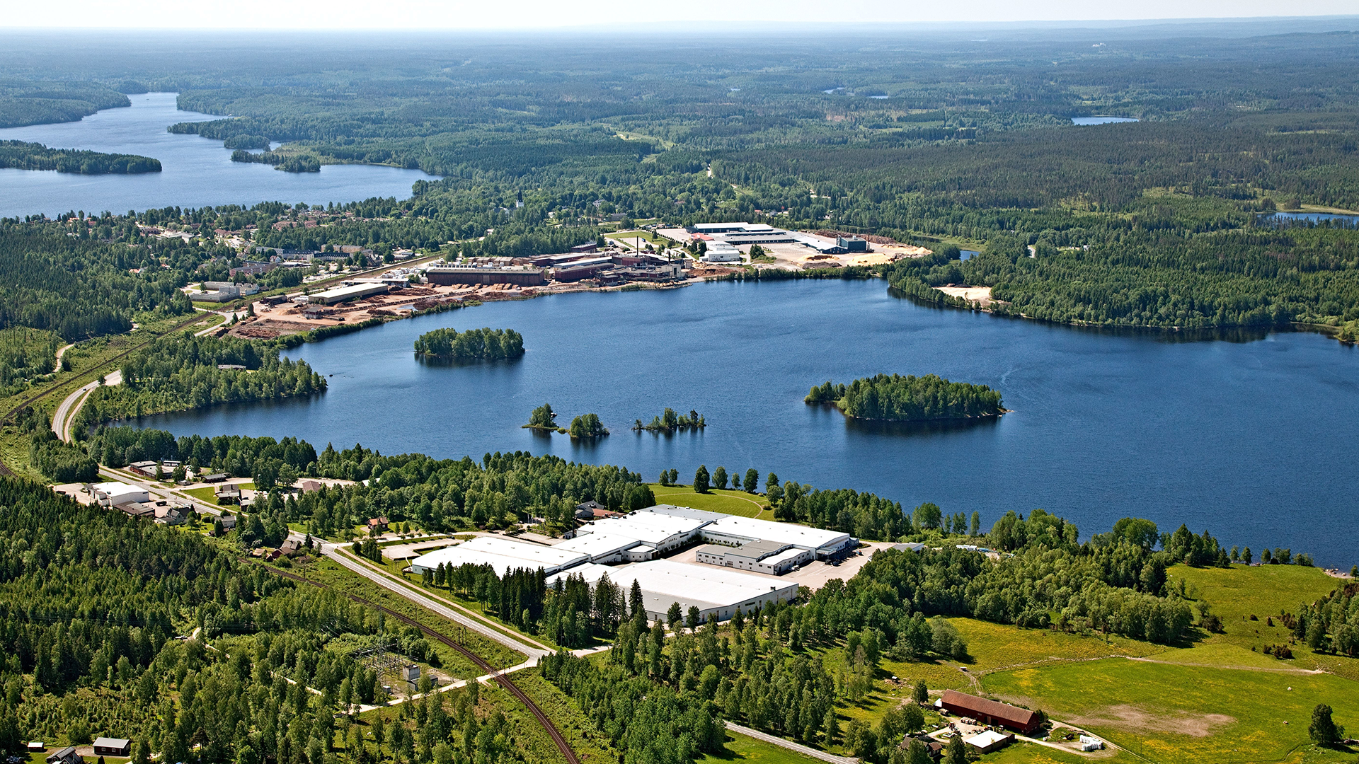 Flygfoto över en industrianläggning vid en stor sjö omgiven av skog och gröna marker.