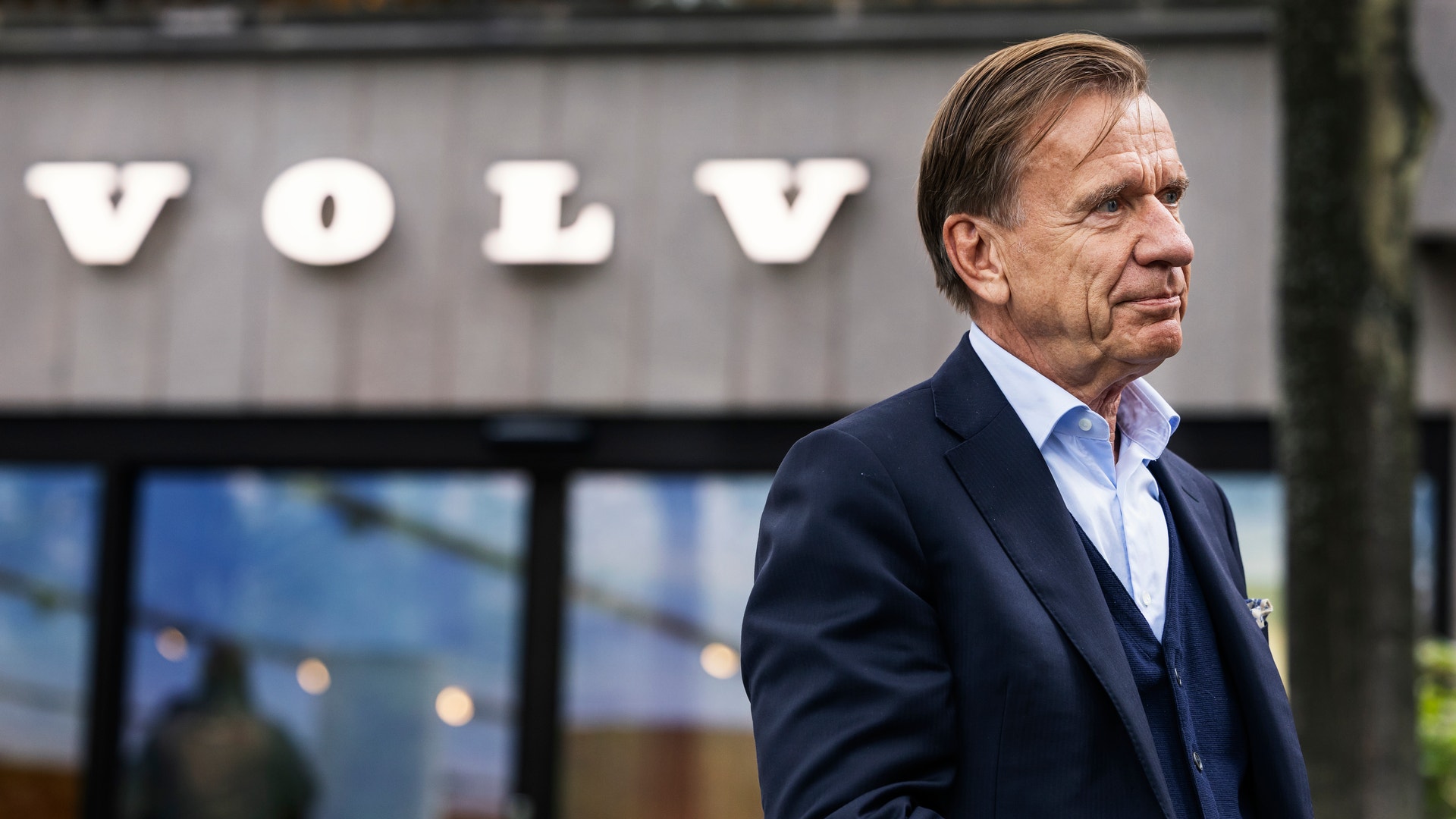 En man i kostym står utomhus framför en byggnad med Volvos logotyp.