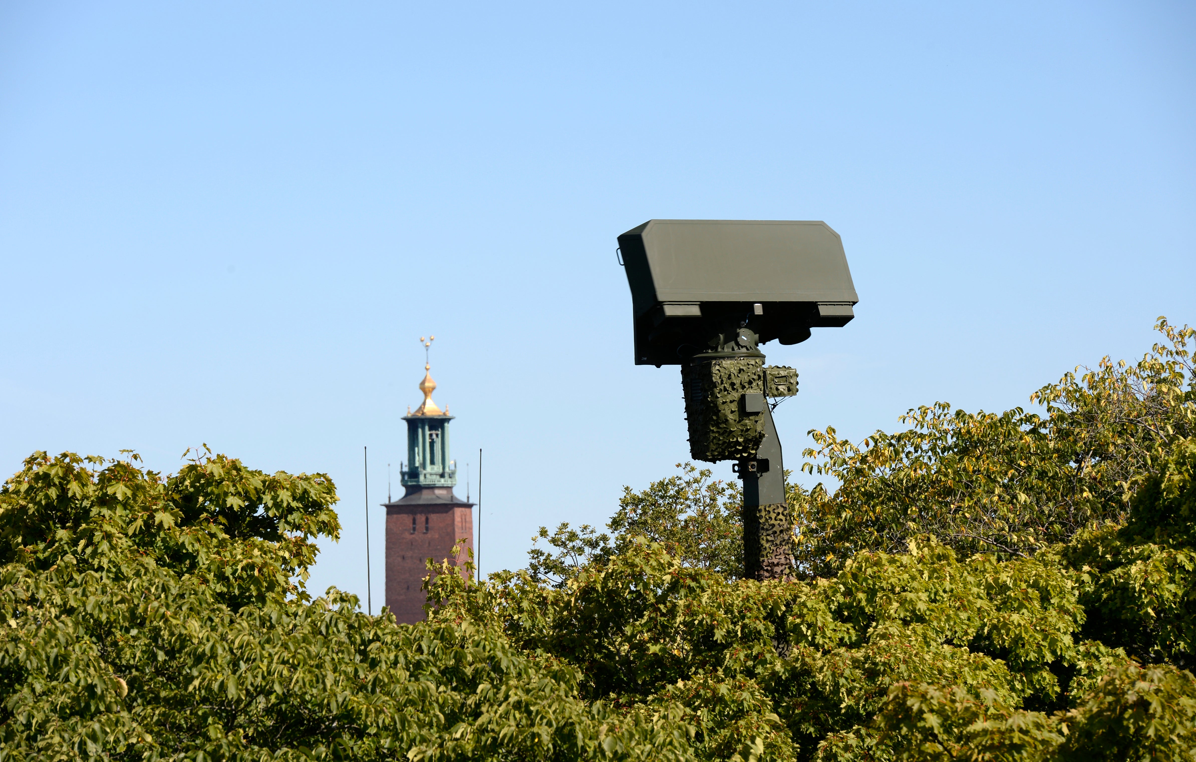 Övervakningskamera monterad på en stolpe bland gröna träd, med ett torn med grön spira synligt i bakgrunden mot blå himmel.