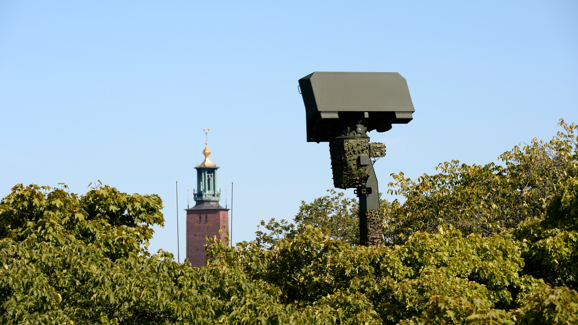Övervakningskamera monterad på en stolpe bland gröna träd, med ett torn med grön spira synligt i bakgrunden mot blå himmel.