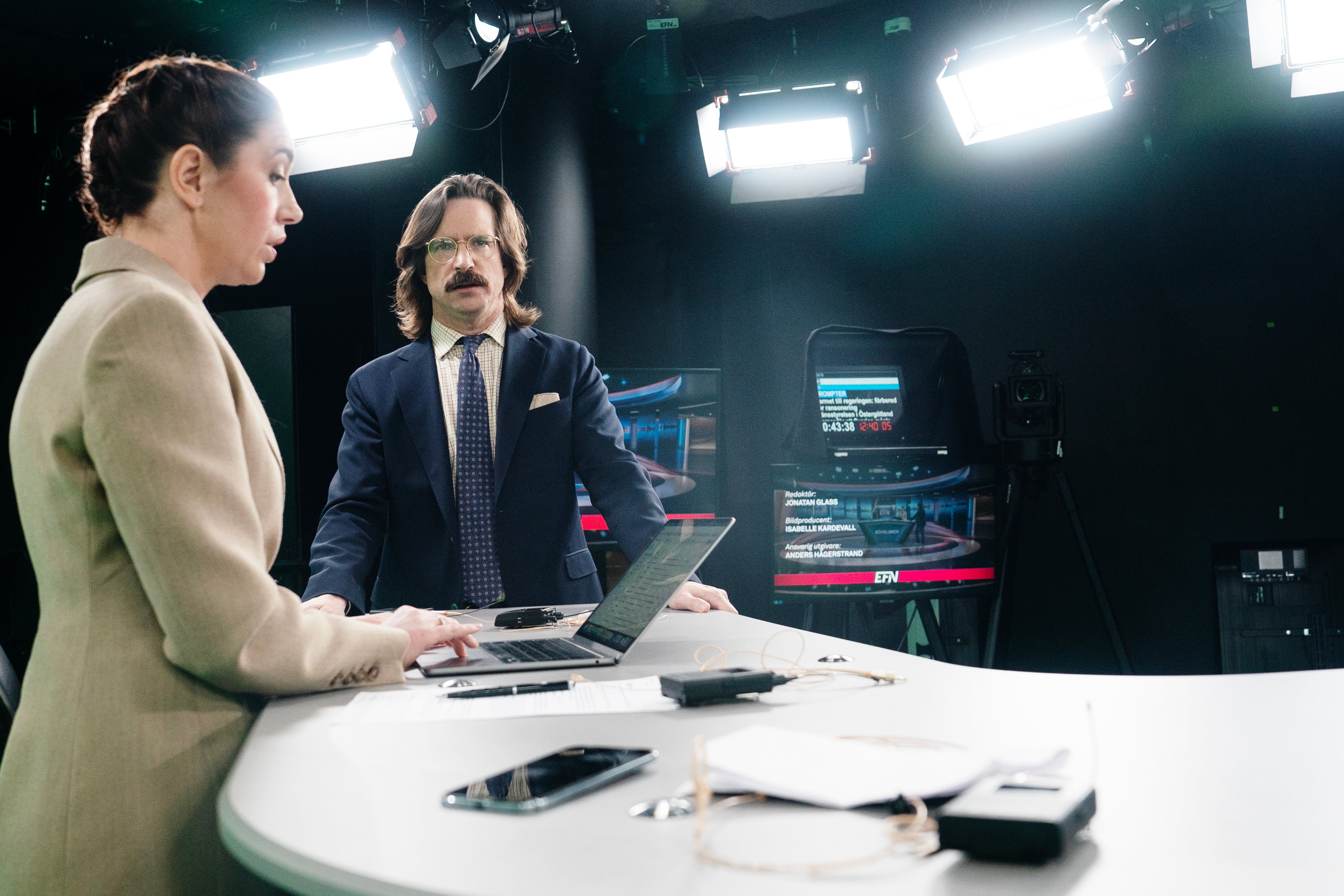 Två personer sitter vid ett skrivbord i en TV-studio och diskuterar framför datorer och studioutrustning.