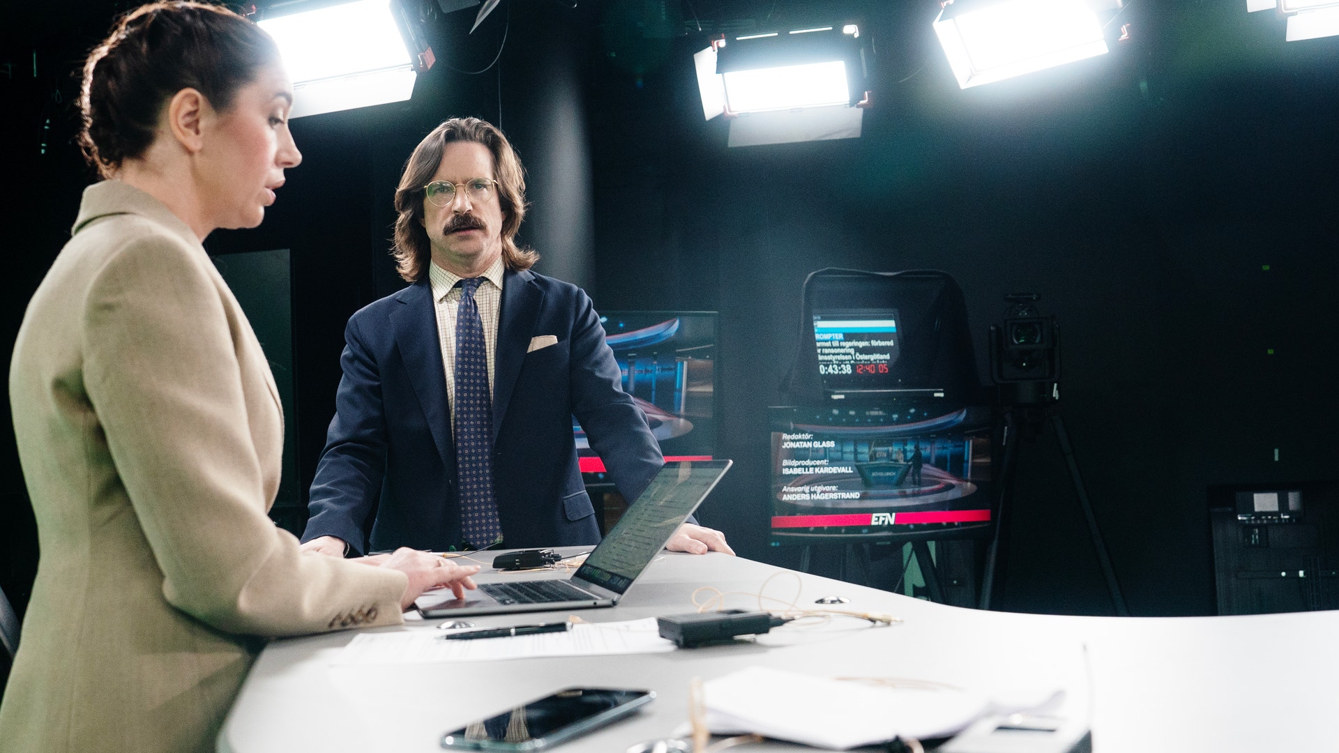 Två personer sitter vid ett skrivbord i en TV-studio och diskuterar framför datorer och studioutrustning.