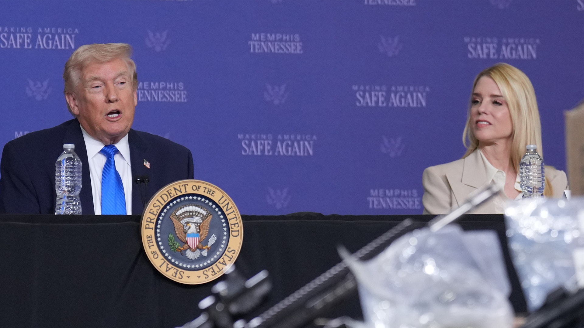 Man talar vid presidentpodium med kvinnlig assistent sittande bredvid honom.