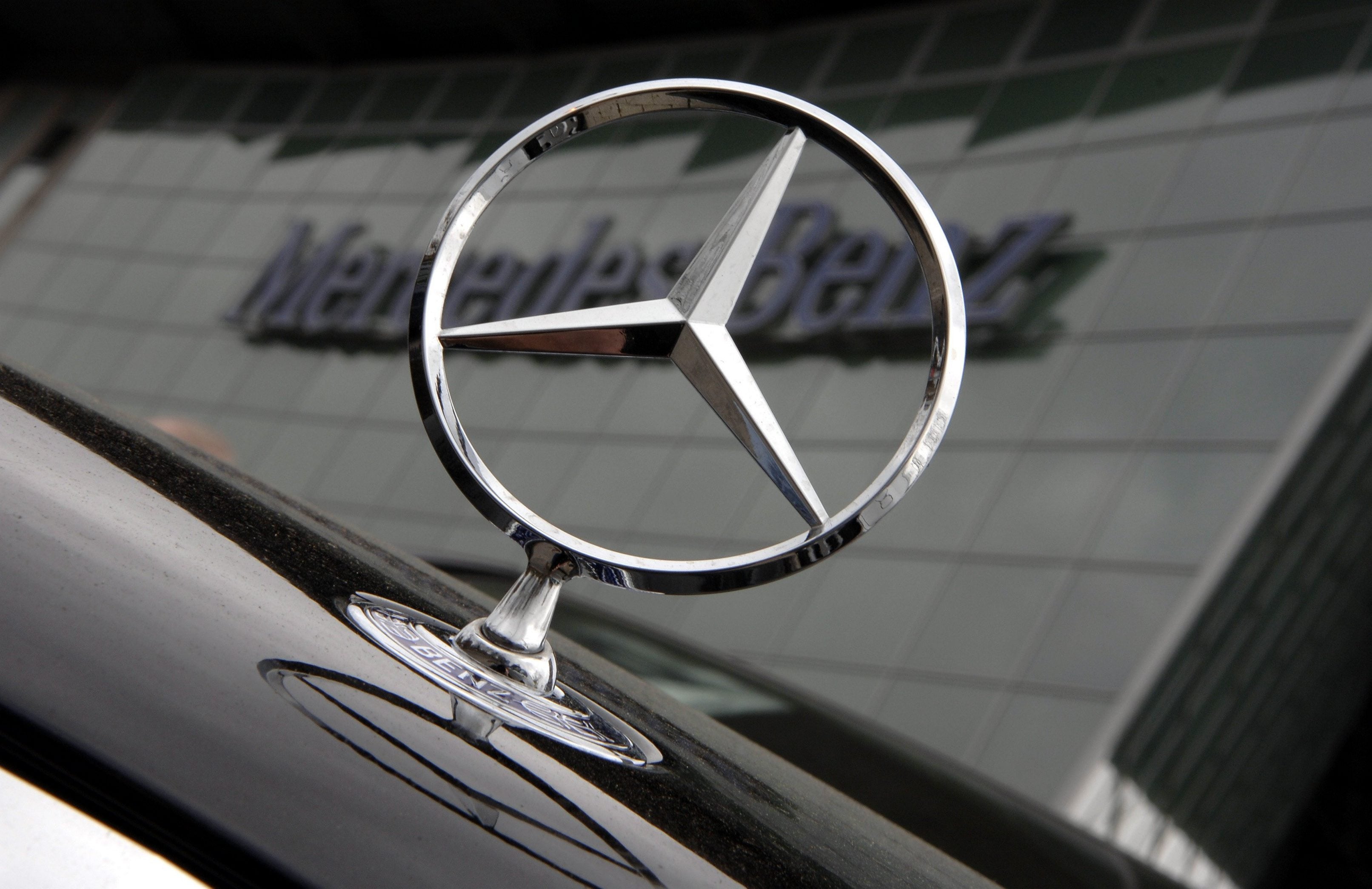 Mercedes-stjärnan på huven av en svart bil, framför en byggnad med Mercedes-Benz-skylt.