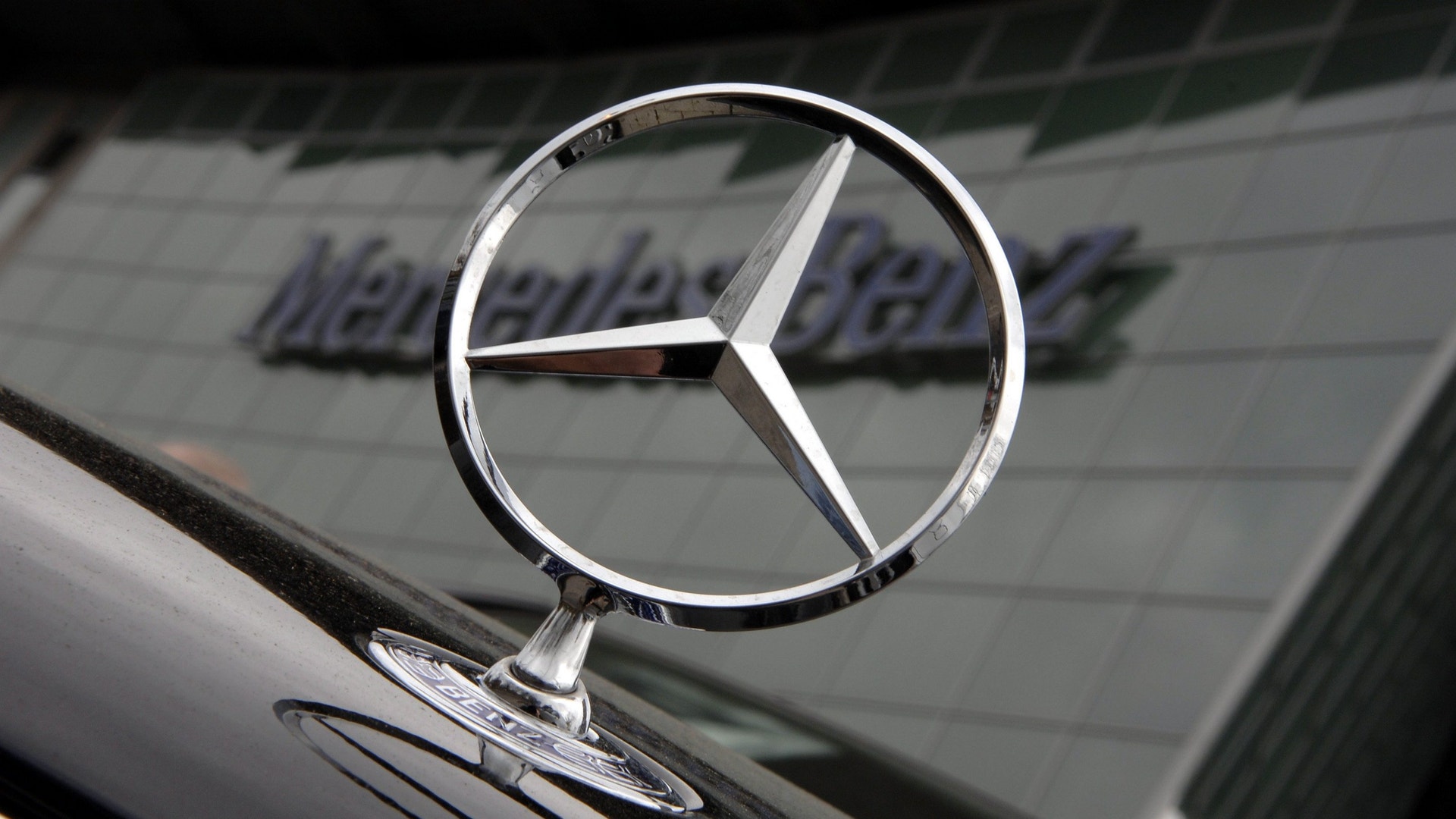 Mercedes-stjärnan på huven av en svart bil, framför en byggnad med Mercedes-Benz-skylt.