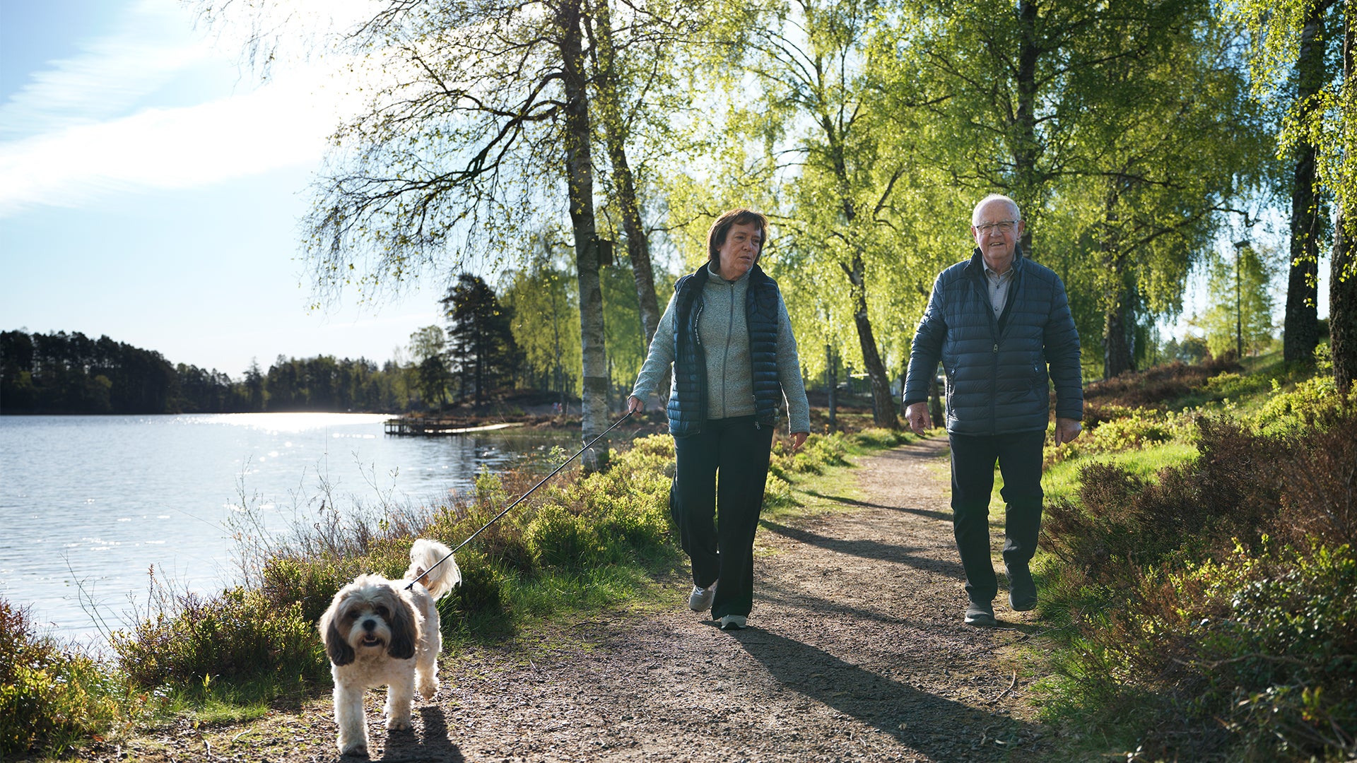 Två personer och en hund går på en stig längs en sjö omgiven av träd och grönska på en solig dag.