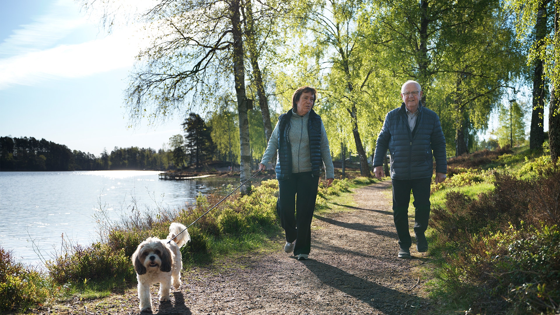 Två personer och en hund går på en stig längs en sjö omgiven av träd och grönska på en solig dag.