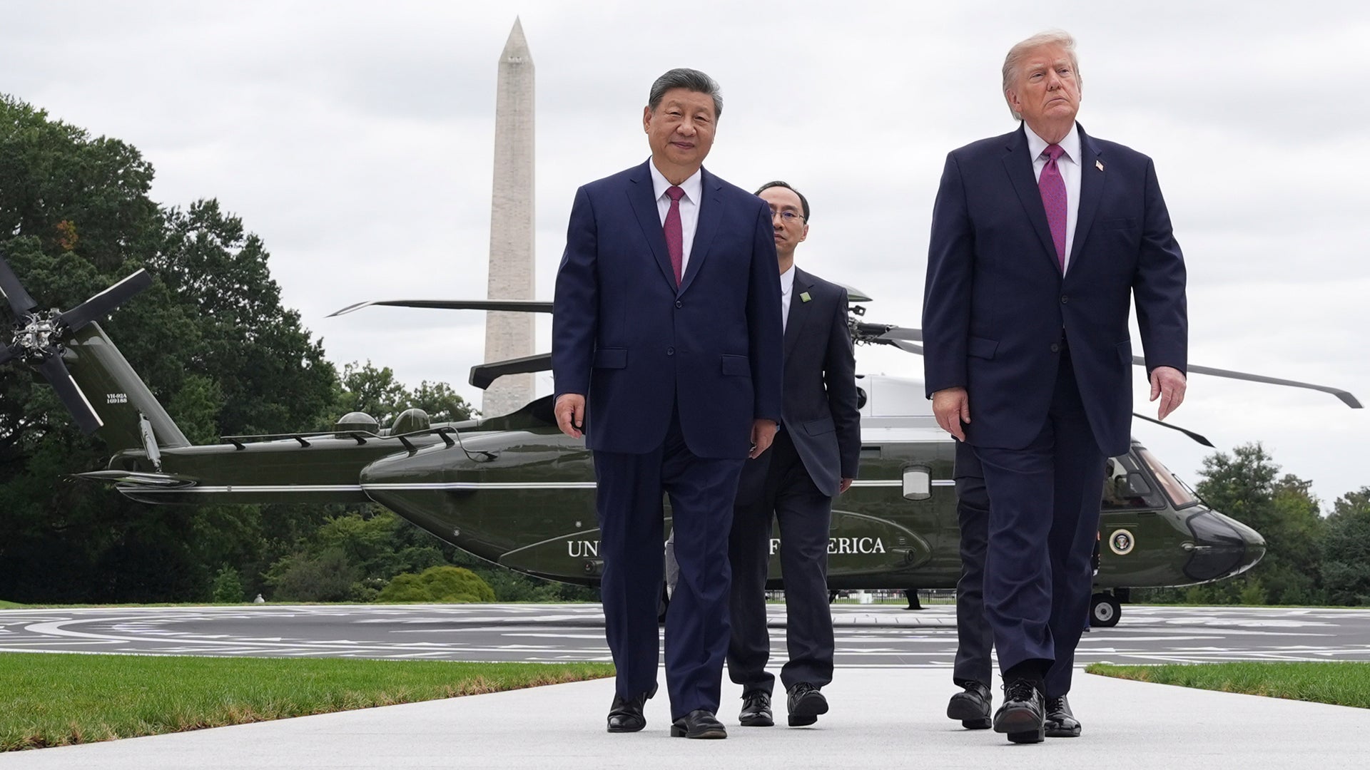 Två män i kostym går tillsammans utomhus med en tolk mellan sig, med helikoptrar och Washington Monument i bakgrunden.