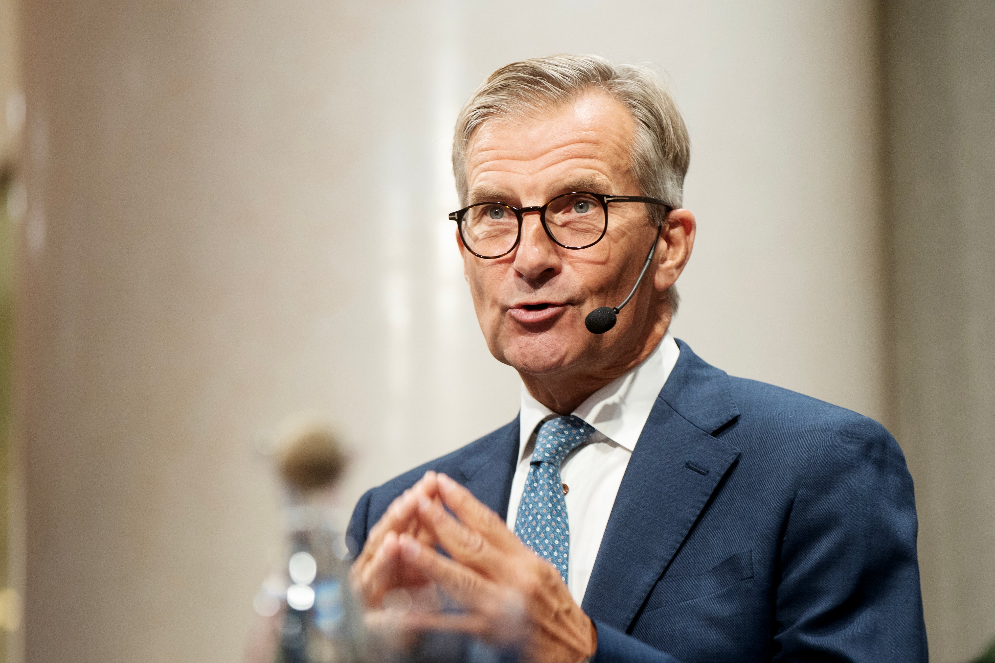 En äldre man i blå kostym och glasögon håller ett föredrag med headset-mikrofon.