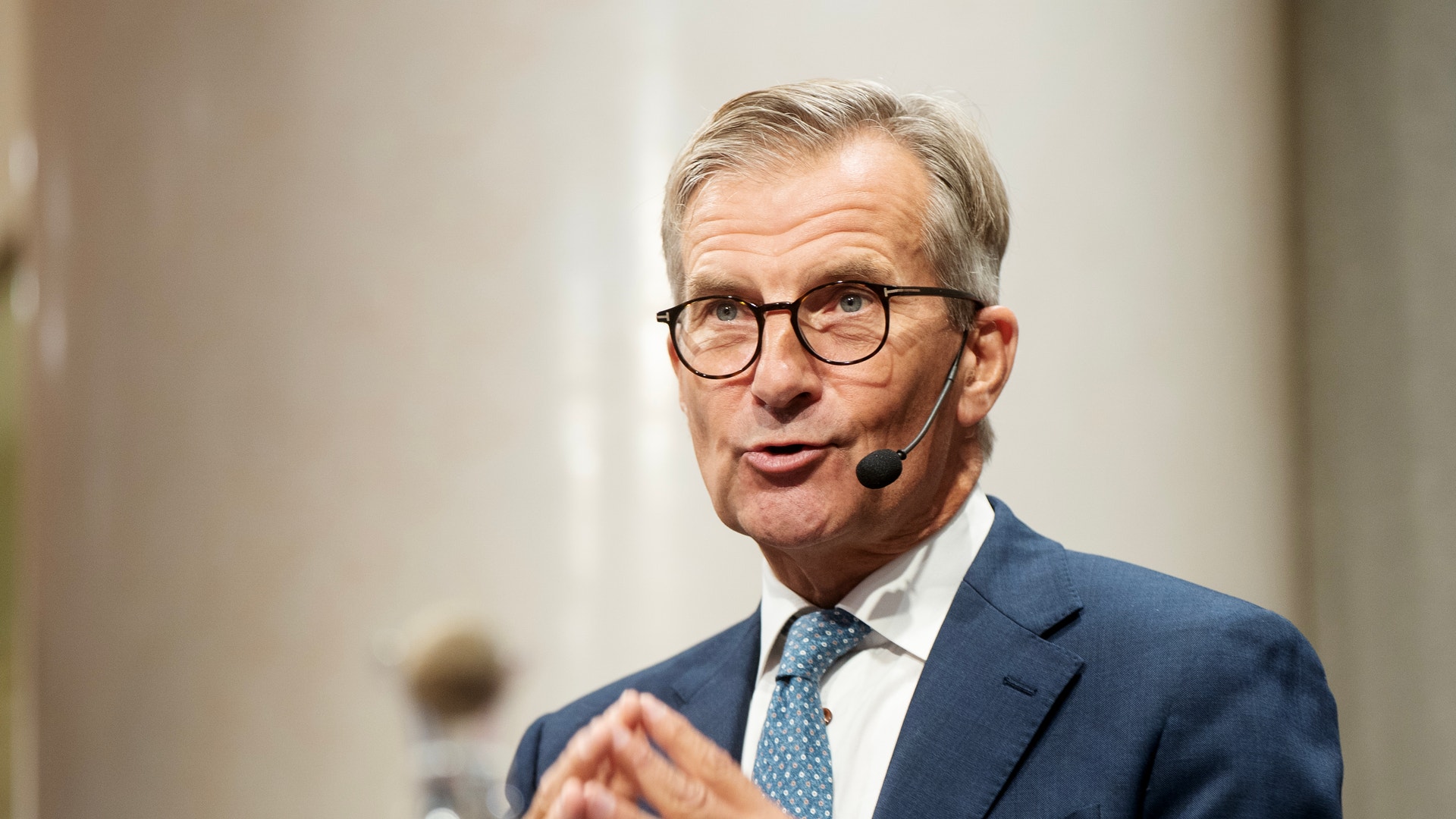 En äldre man i blå kostym och glasögon håller ett föredrag med headset-mikrofon.
