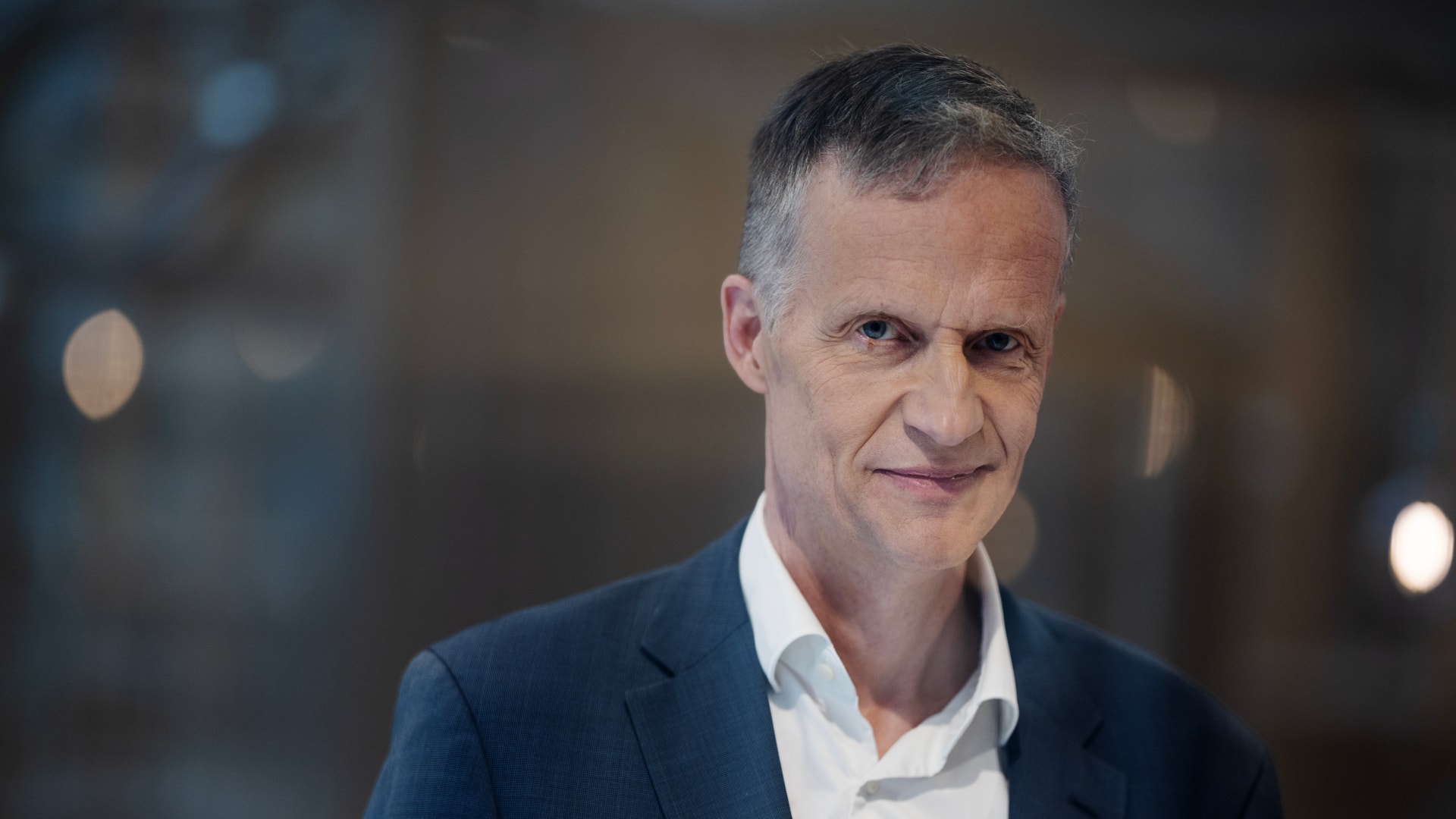 Porträtt av en man i mörkblå kavaj och vit skjorta mot en suddig bakgrund.