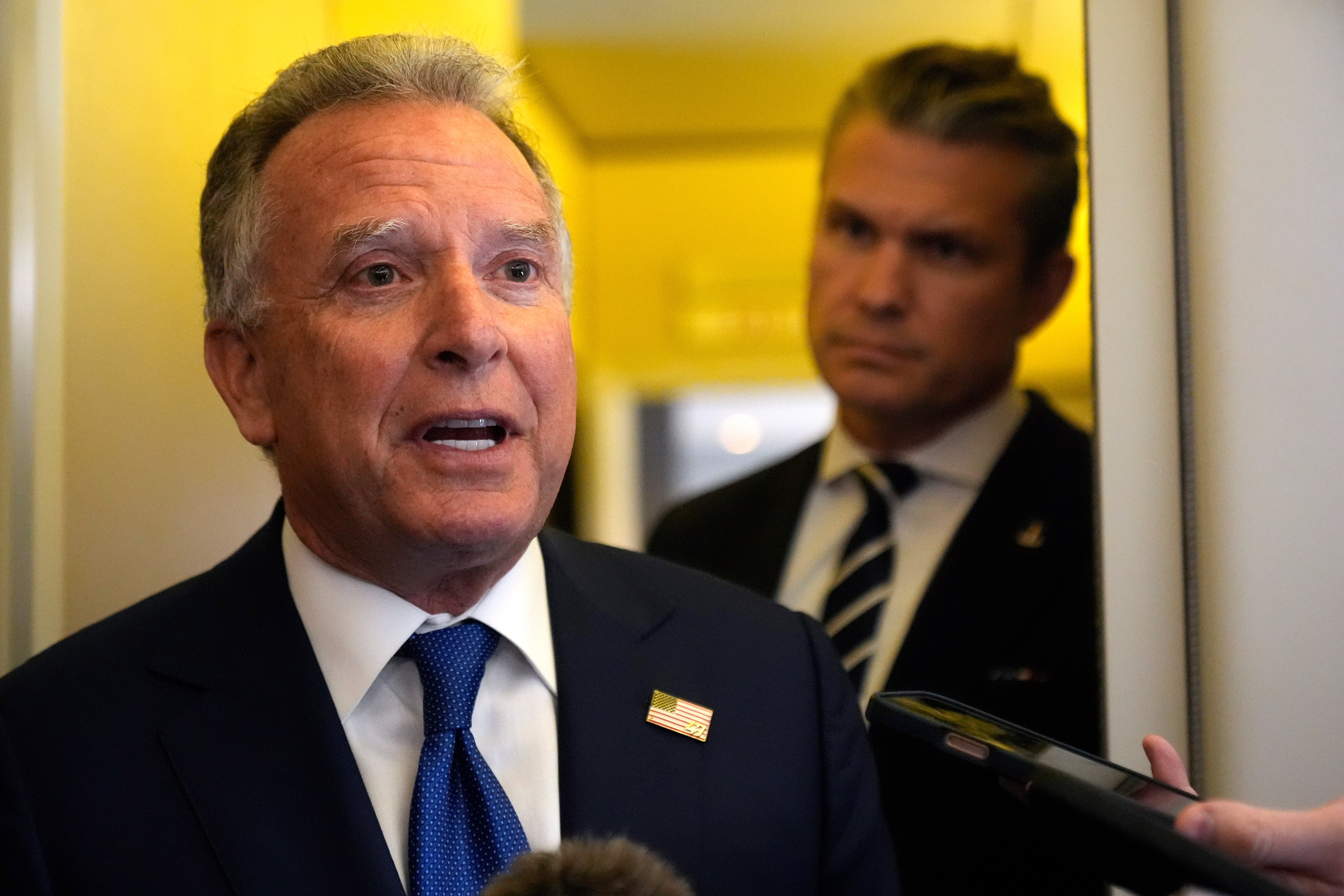 En man i kostym och blå slips talar vid en mikrofon i en professionell miljö.