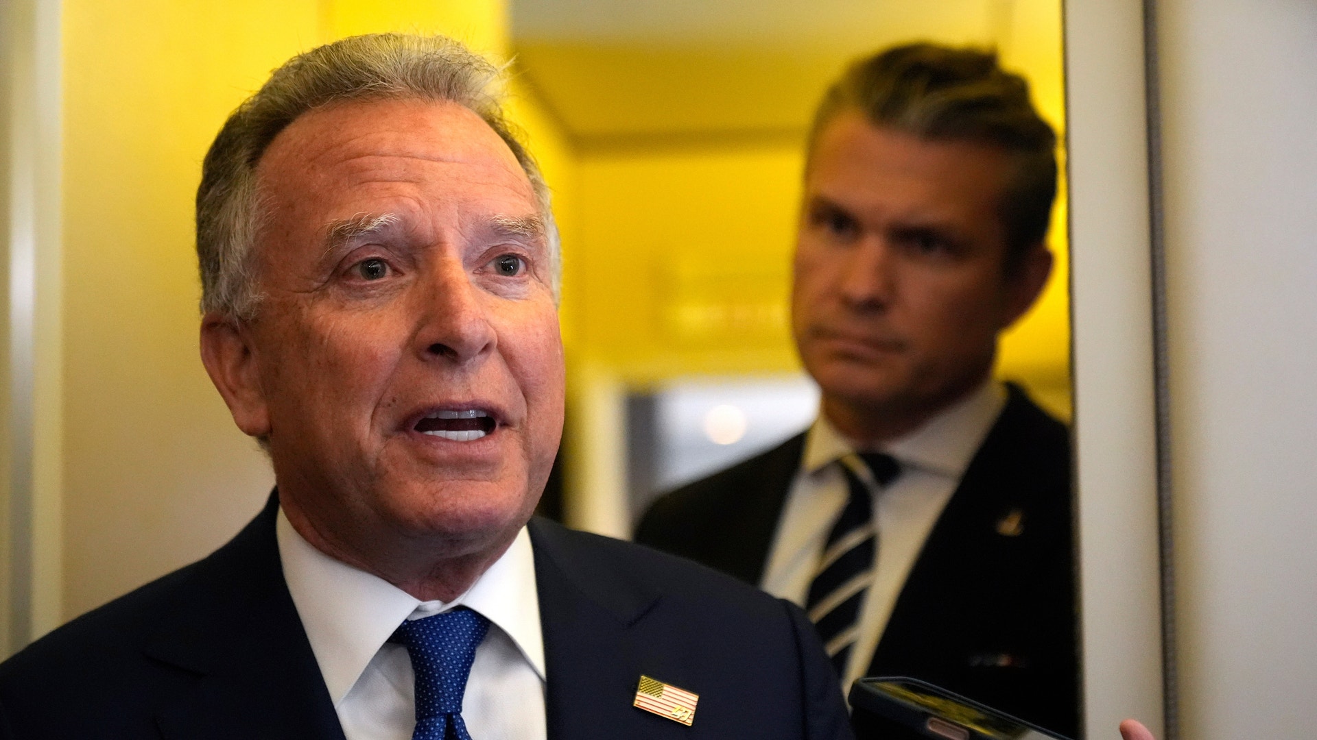 En man i kostym och blå slips talar vid en mikrofon i en professionell miljö.