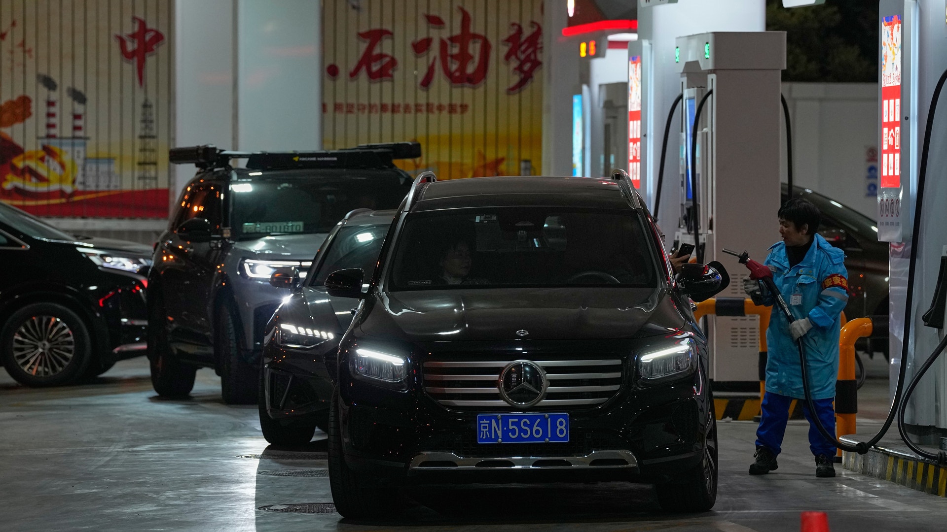 Bilar vid en bensinstation på kvällen, en tankkassörskan fyller på en svart bil.