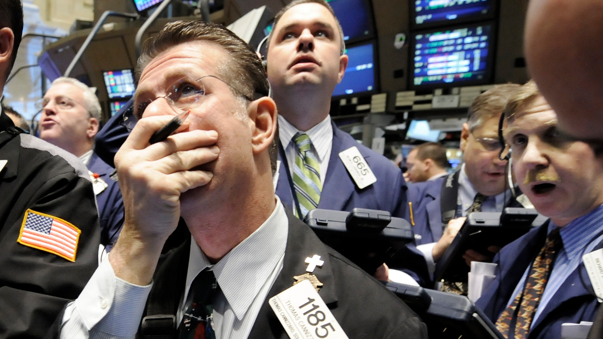 Handlare på en aktiebörs med bekymrad uppsyn, omgivna av kolleger och handelsskärmar i bakgrunden.
