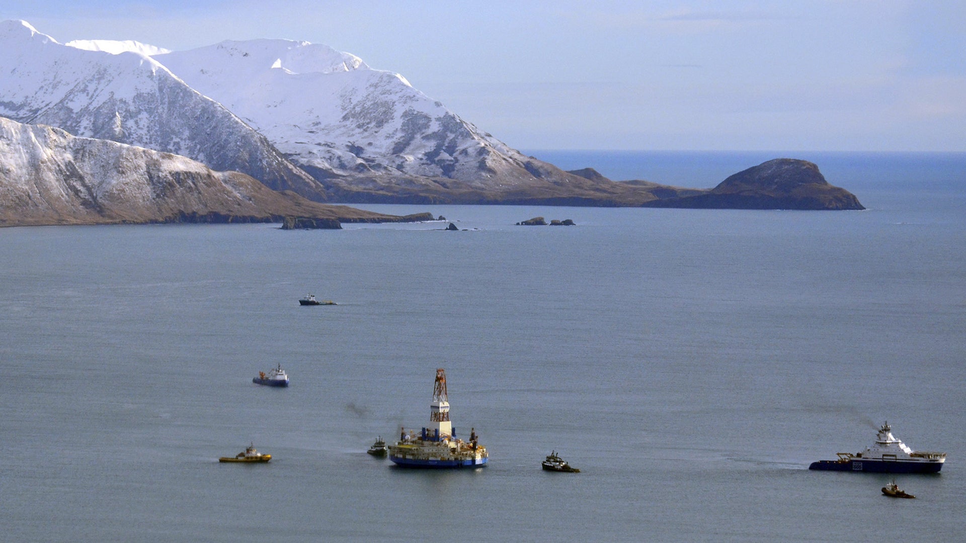 En oljeborrplattform bogseras av fartyg genom en fjord med snötäckta berg i bakgrunden.