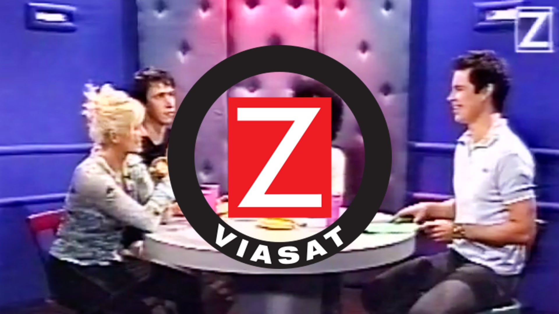 TV-studio med tre personer sittande vid ett bord med Z Viasat-logon i förgrunden.