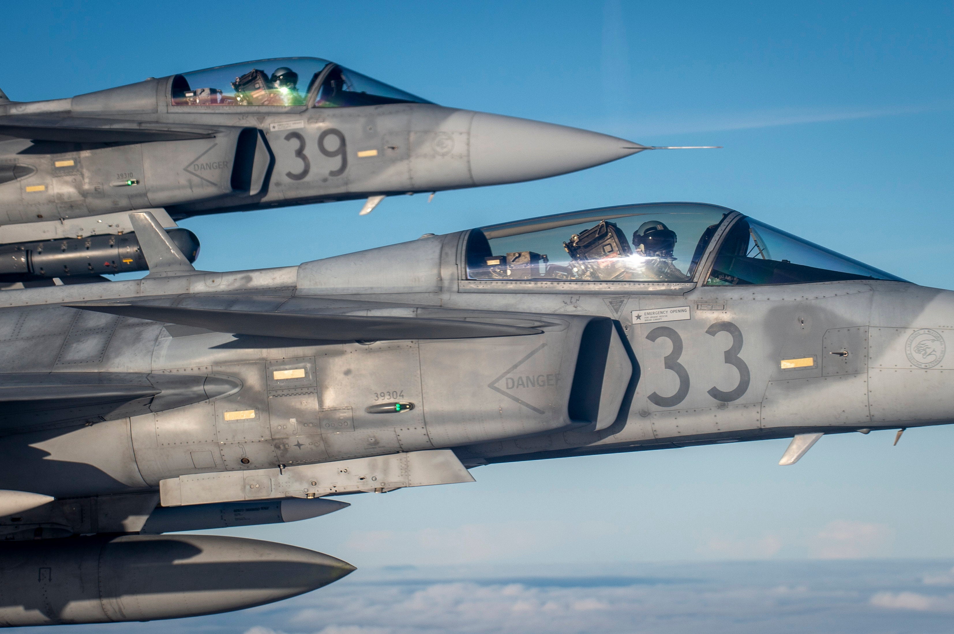 Två stridsflygplan fotograferade från luften mot blå himmel. Piloterna syns i cockpitens glasskupor. Flygplanen har nummer på sidorna.