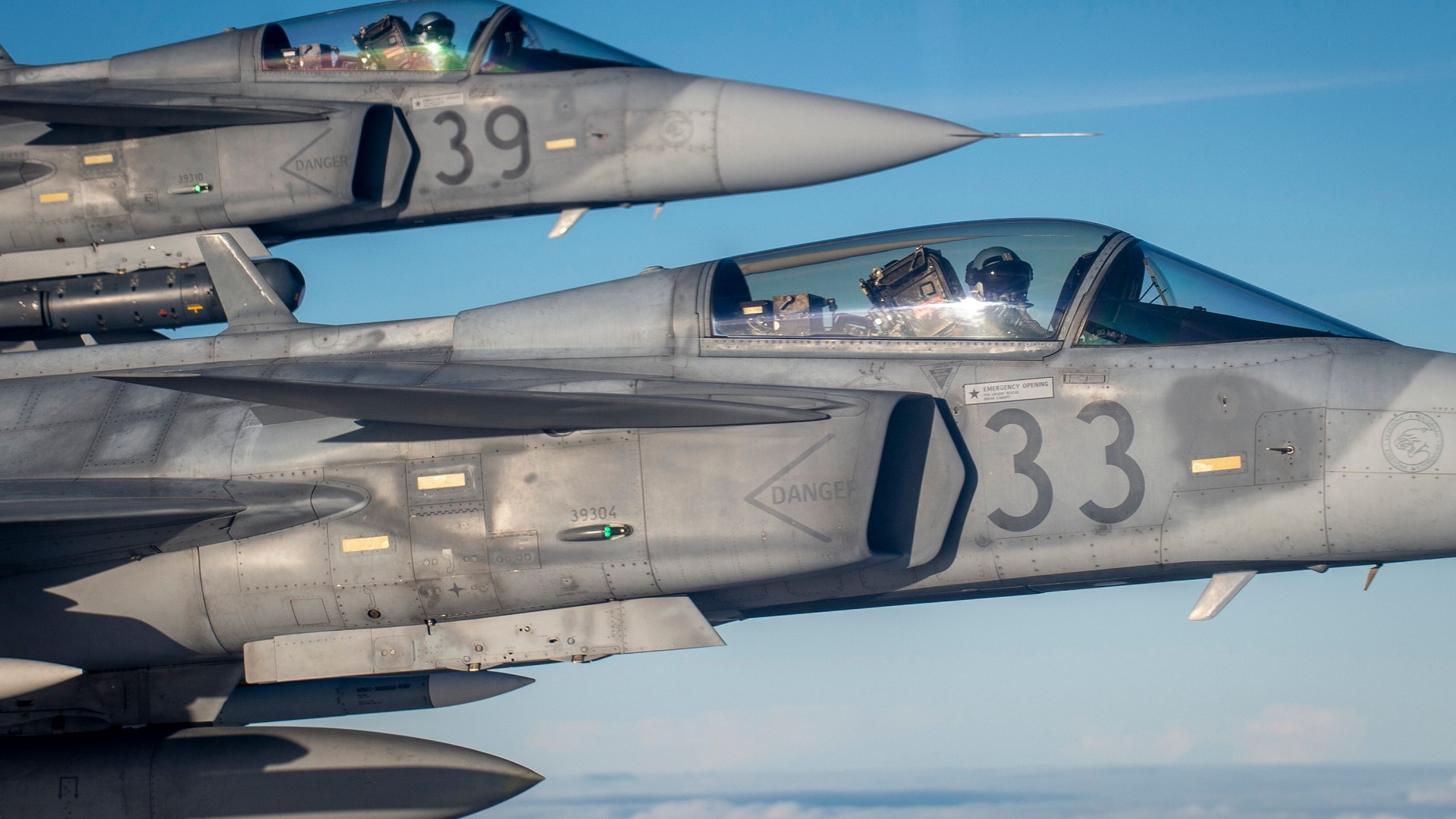 Två stridsflygplan fotograferade från luften mot blå himmel. Piloterna syns i cockpitens glasskupor. Flygplanen har nummer på sidorna.