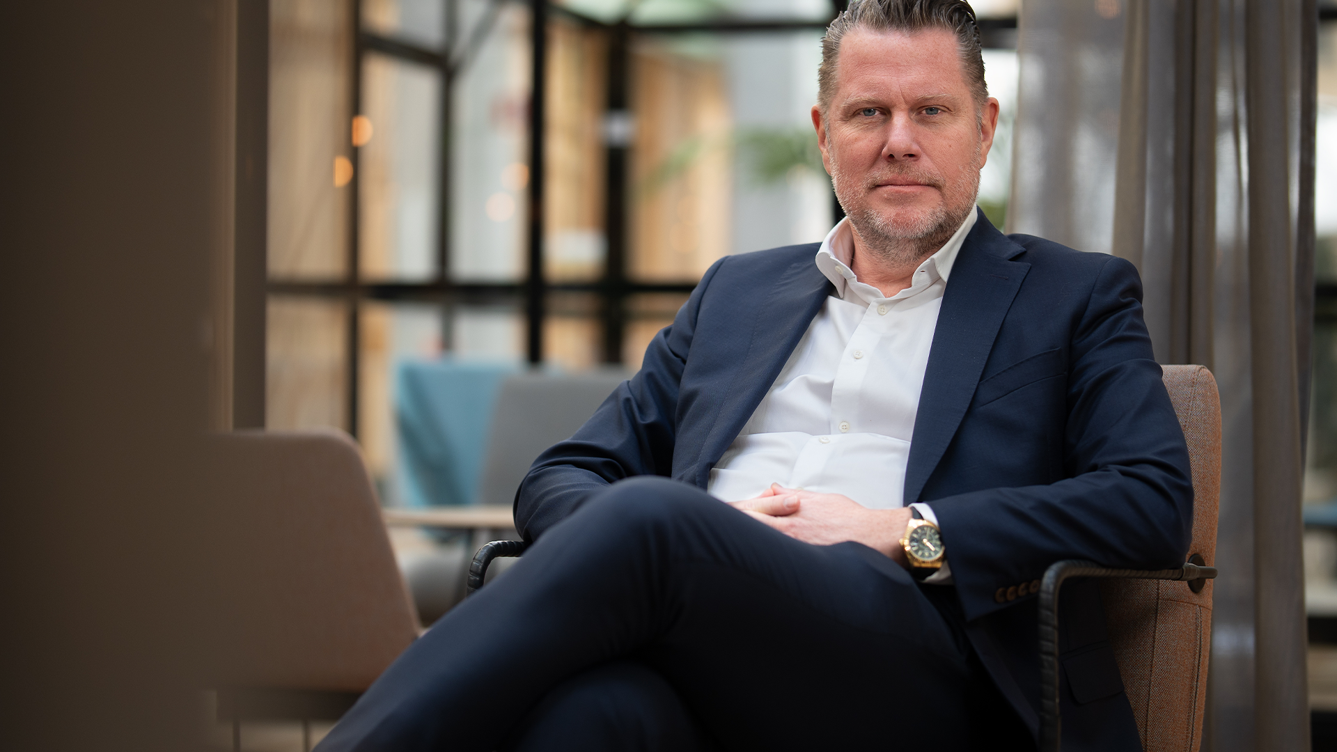 Porträtt av en man i mörkblå kostym och vit skjorta som sitter i en stol i en modern kontorsmiljö.