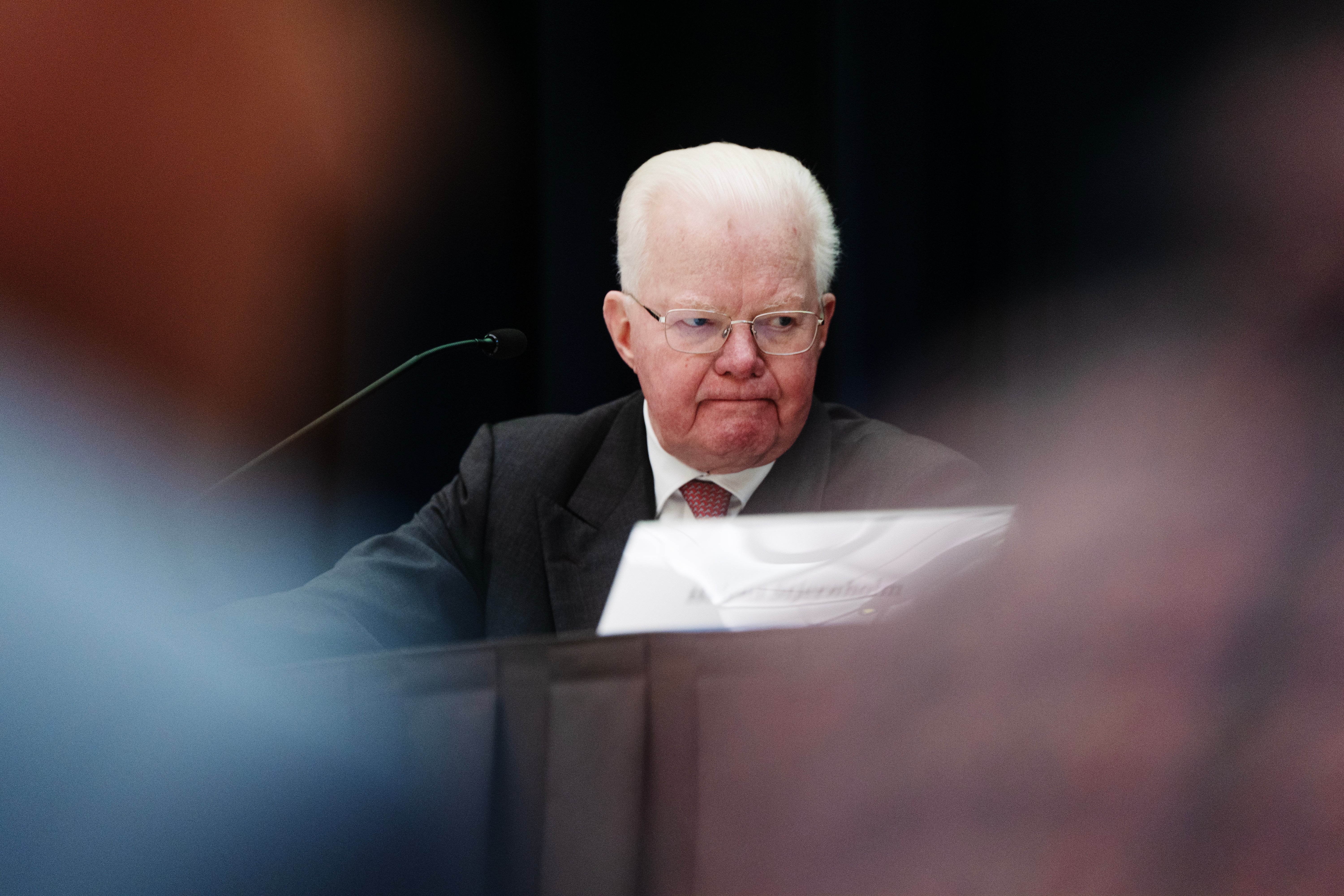 En äldre man i kostym och glasögon talar vid ett podium med mikrofon framför en suddig publik.